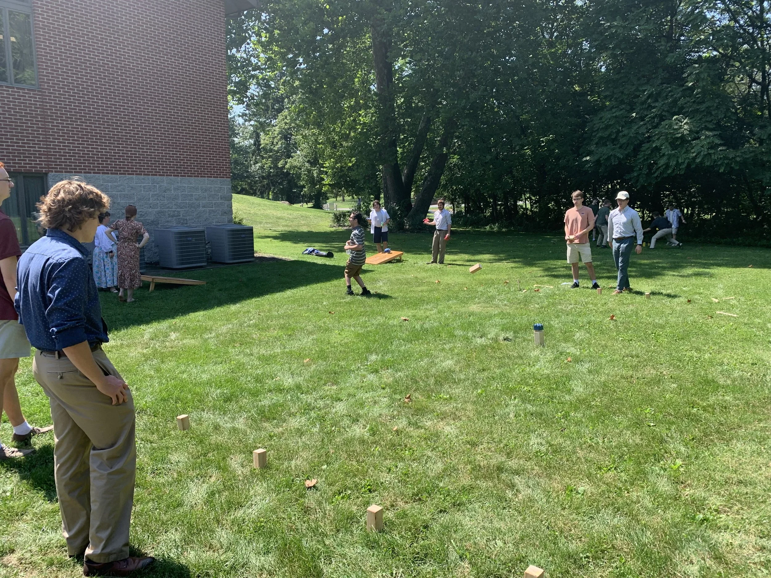 People playing a game of cornhole on a grassy lawn in sunny weather, with some spectators watching.