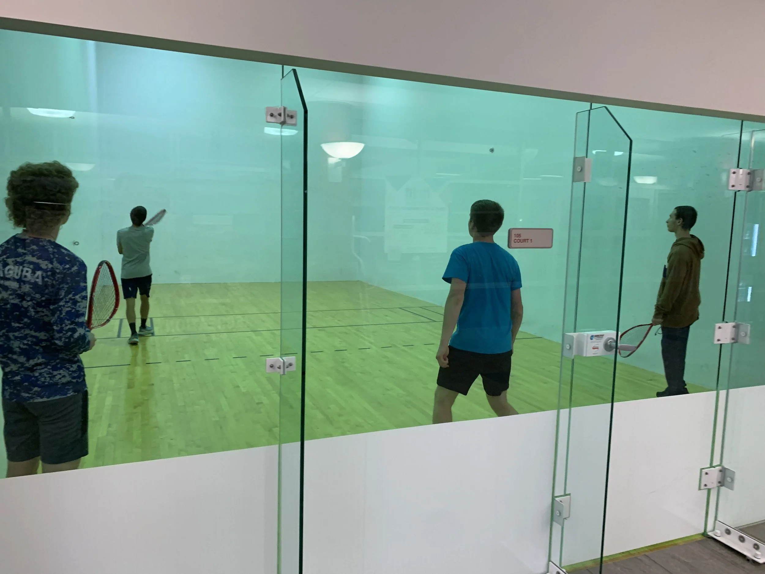 Four young men playing squash inside a glass-walled court at an indoor sports facility. One is preparing to hit a ball, while the others watch.