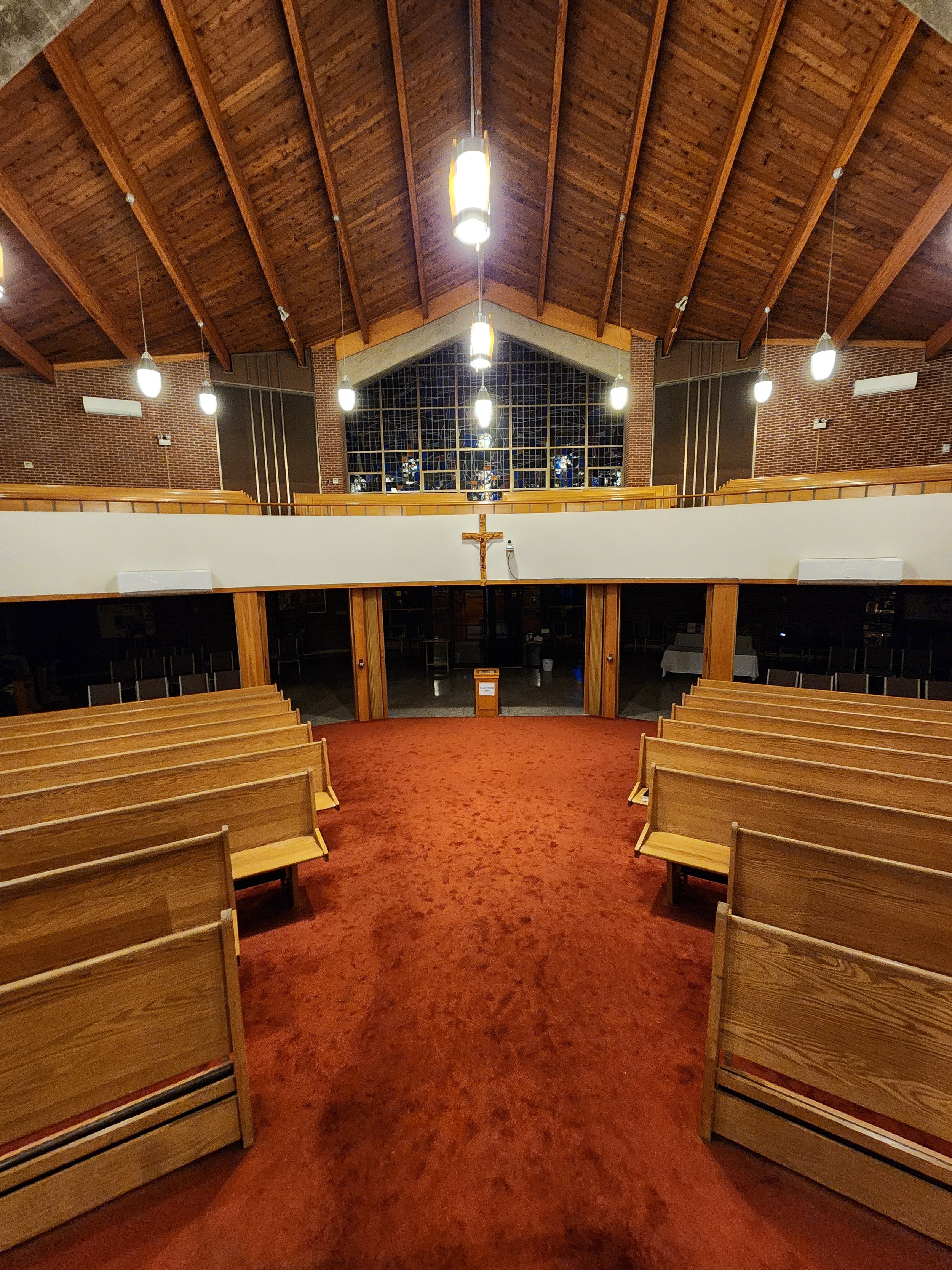 Interior of a church with wooden pews, red carpet, brick walls, a crucifix on the wall, and large stained glass window at the front.