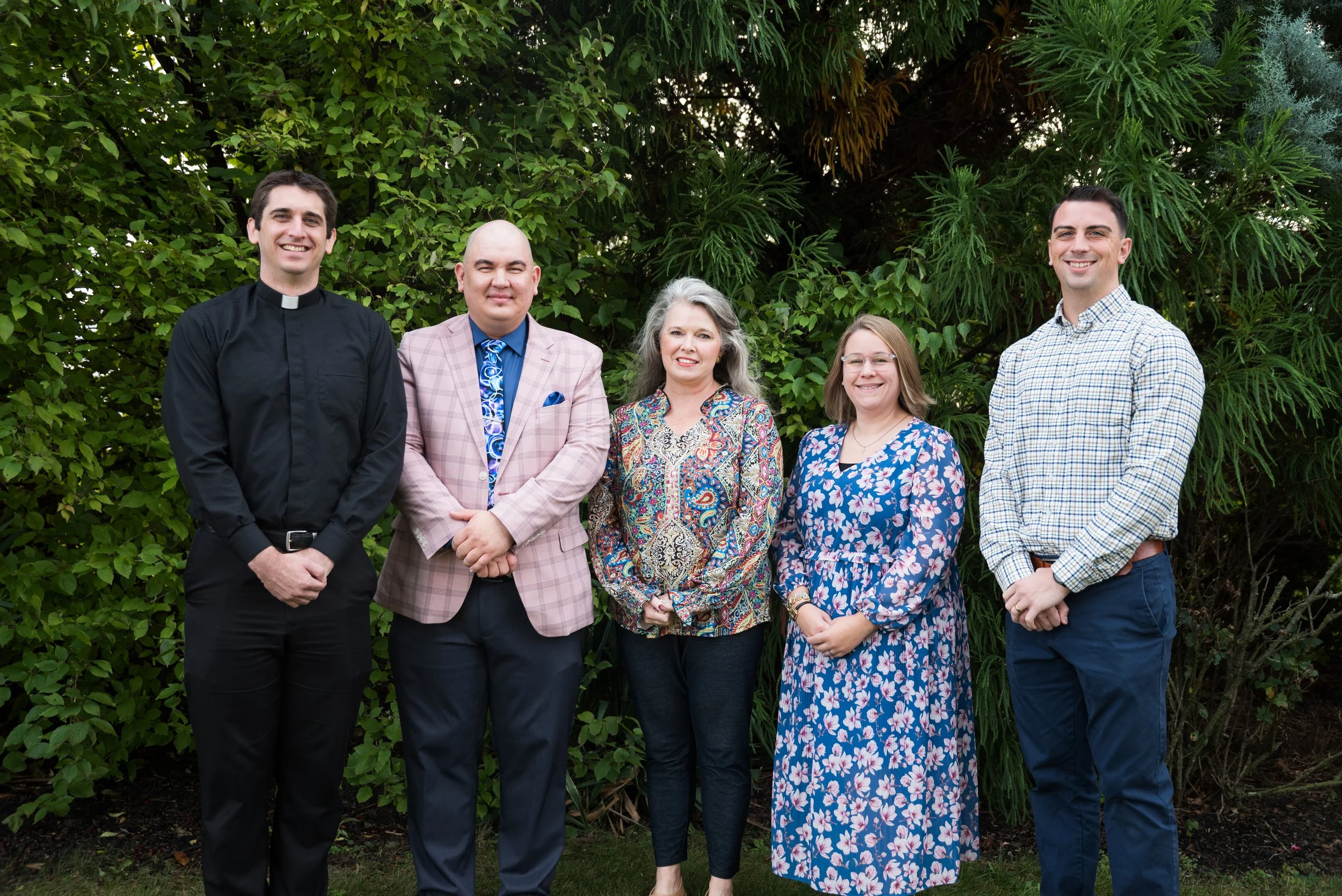 Group of five people standing outdoors in front of green bushes, including a man in a black clergy shirt, a man in a light pink plaid blazer, a woman with gray hair in a colorful blouse, a woman in a floral dress, and a man in a plaid shirt.