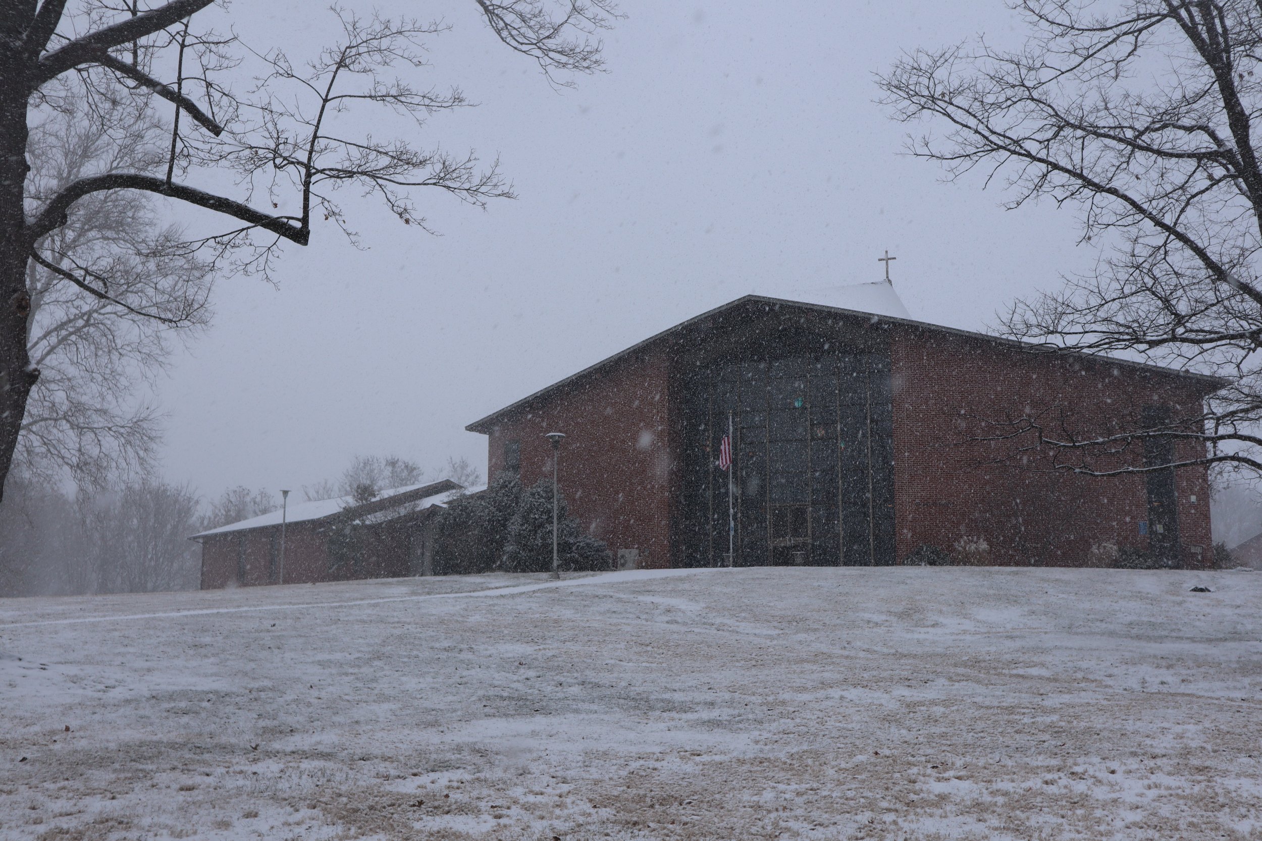 A brick church with a cross on the roof during a snowy day, with leafless trees and snow-covered ground.