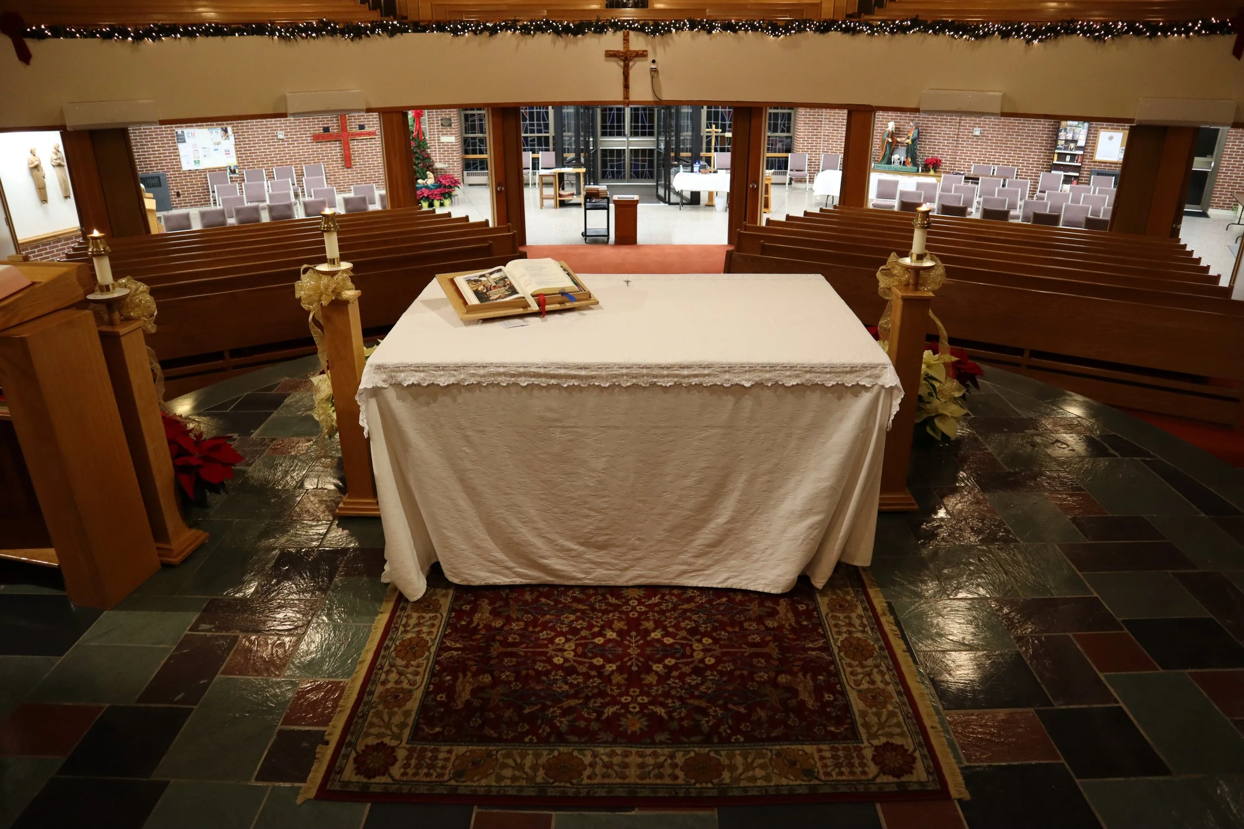 Interior view of a church altar with a white cloth, an open Bible, and Christmas decorations, with pews and stained glass windows in the background.