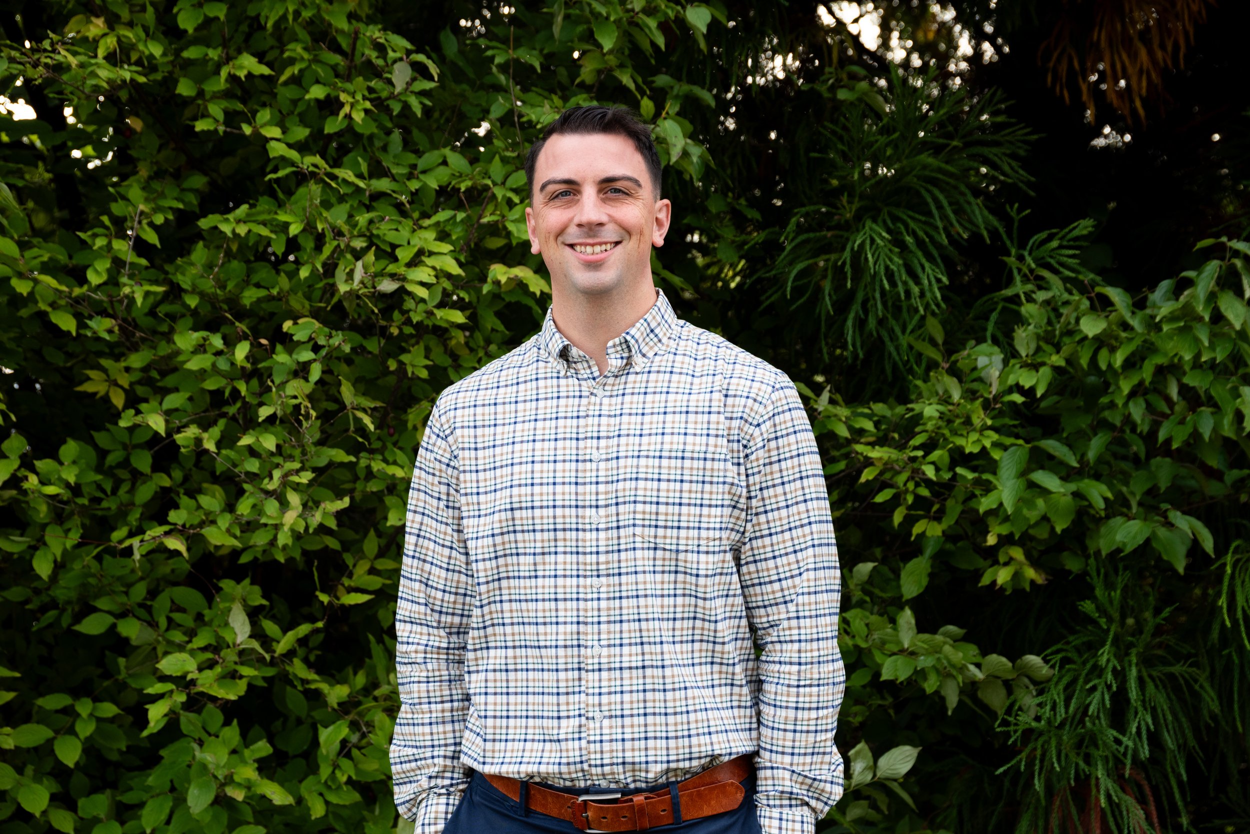 A smiling young man in a plaid shirt standing outdoors in front of green foliage.