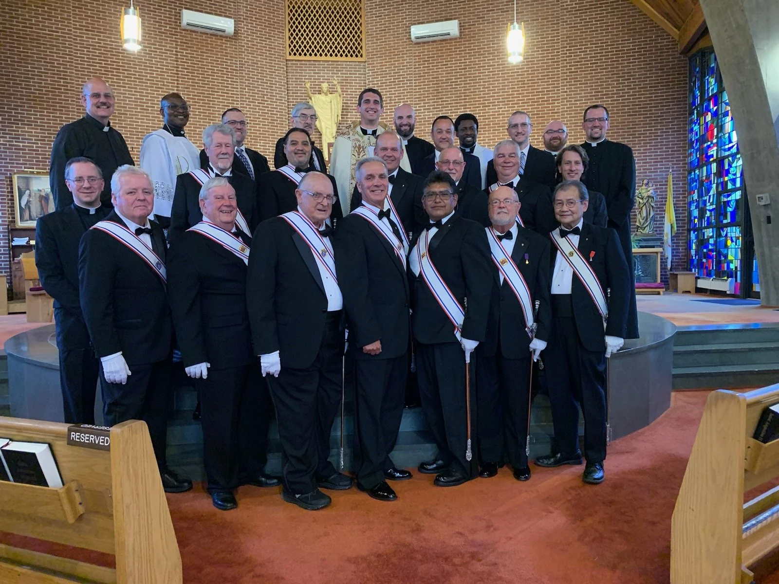 Group of men dressed in formal black tuxedos and robes with sashes, standing inside a church with brick walls, stained glass windows, and religious statues.