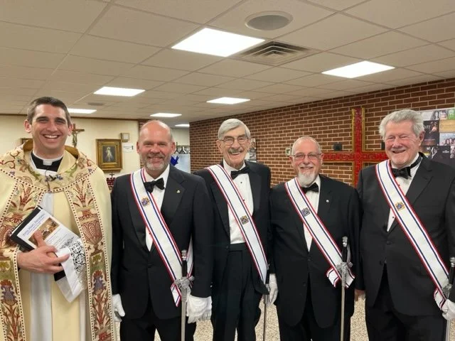 Five men standing inside a church, four wearing tuxedos with sashes, one dressed as a priest or bishop, all smiling and posing for a photo. In the background, a large red cross on the wall.