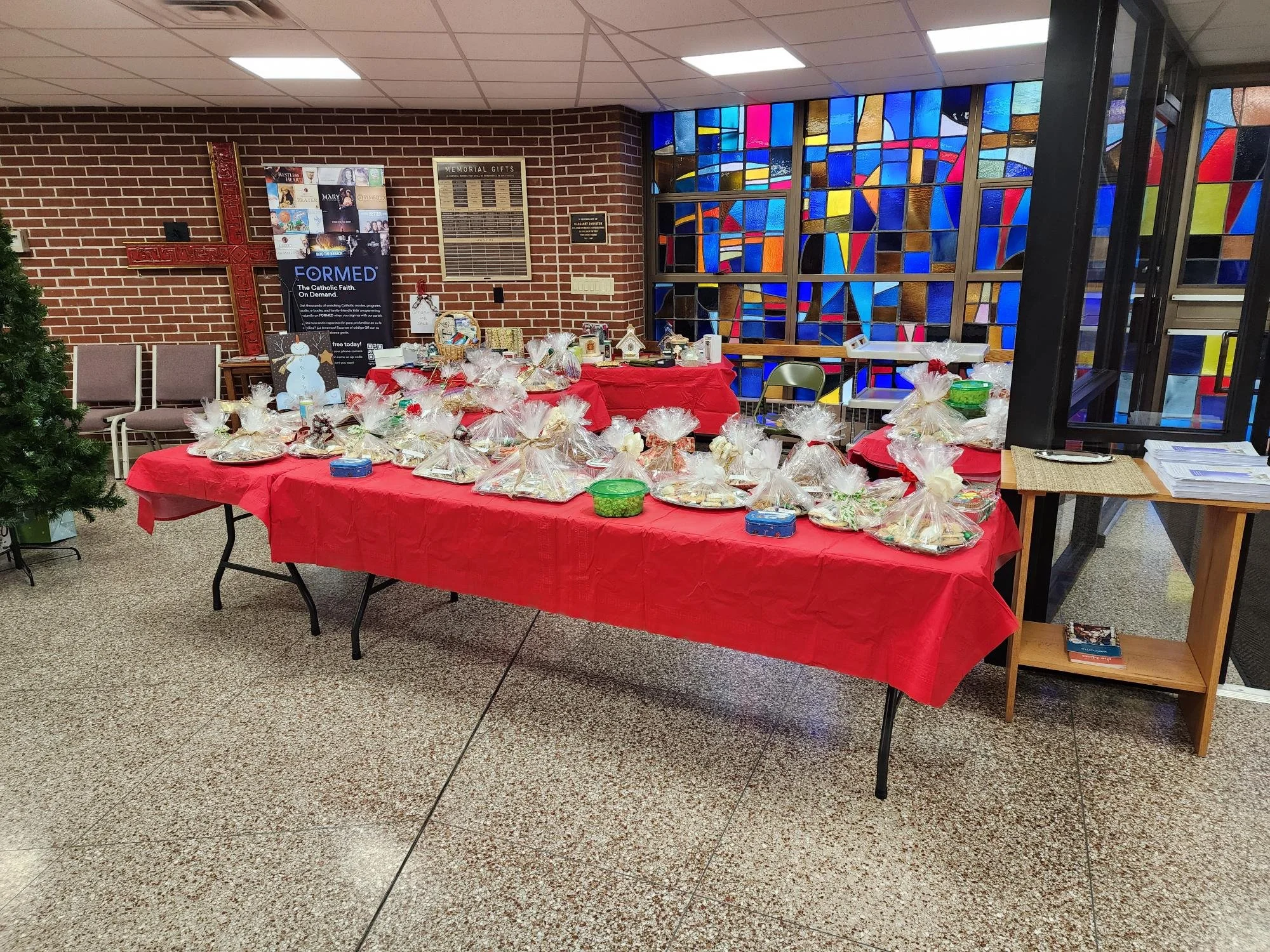 Table with wrapped baked goods and treats, decorated with Christmas-themed items, set up in a church or community center with stained glass windows and brick walls.