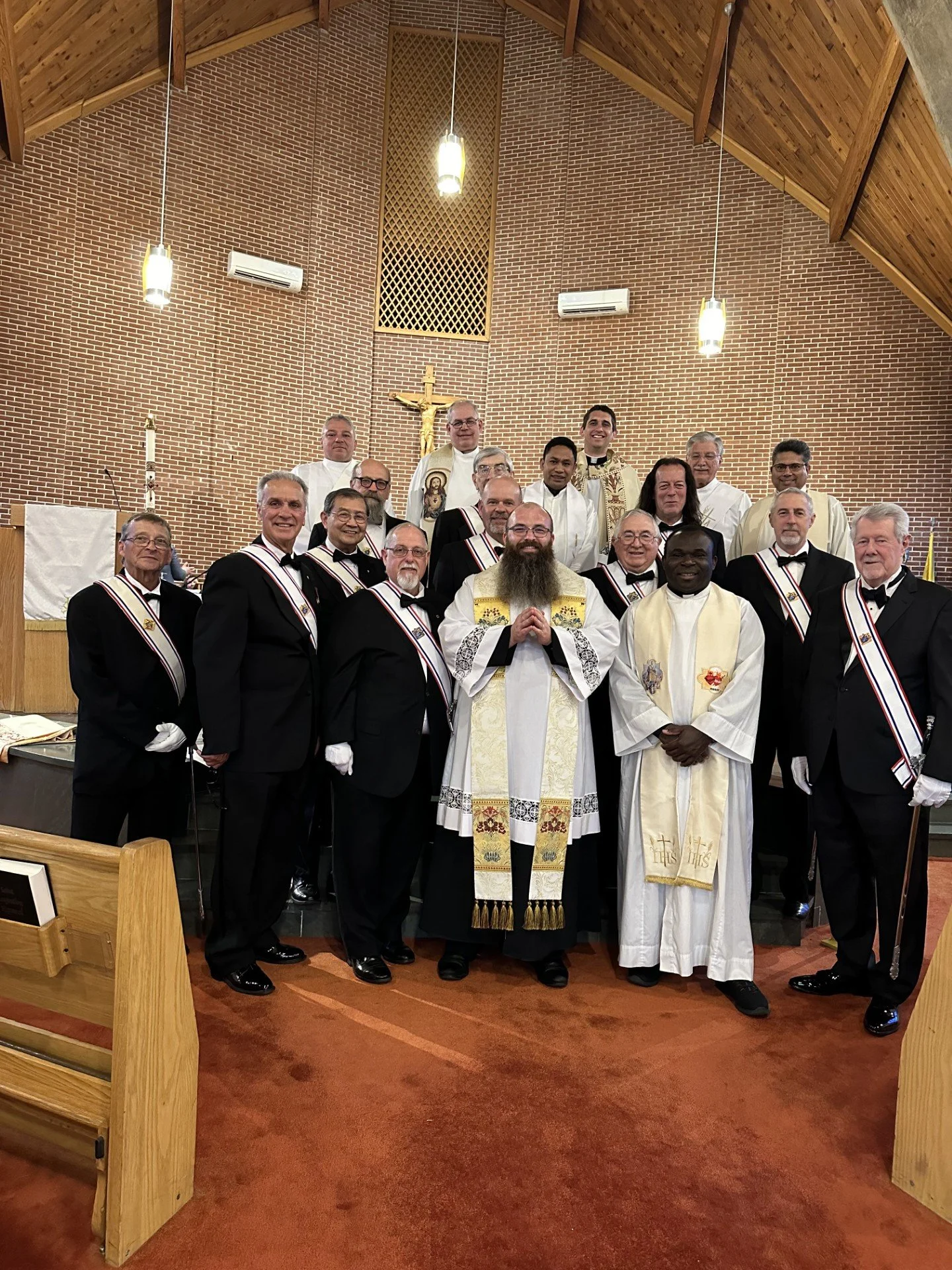 A group of clergy and choir members in church, dressed in formal attire, standing on church altar. Background includes altar with a crucifix, brick wall, and church ceiling.