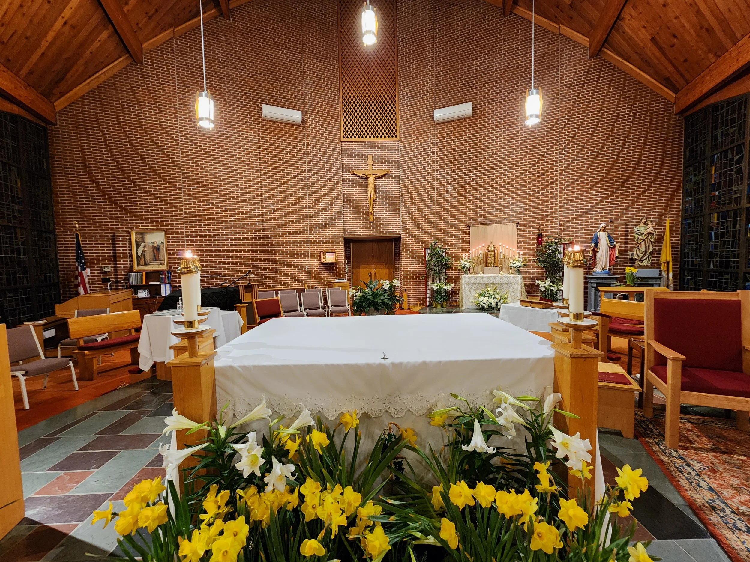 Interior of a church altar decorated with white and yellow flowers, candles, and religious statues, with wooden pews and chairs, a crucifix on a brick wall, and stained glass windows.