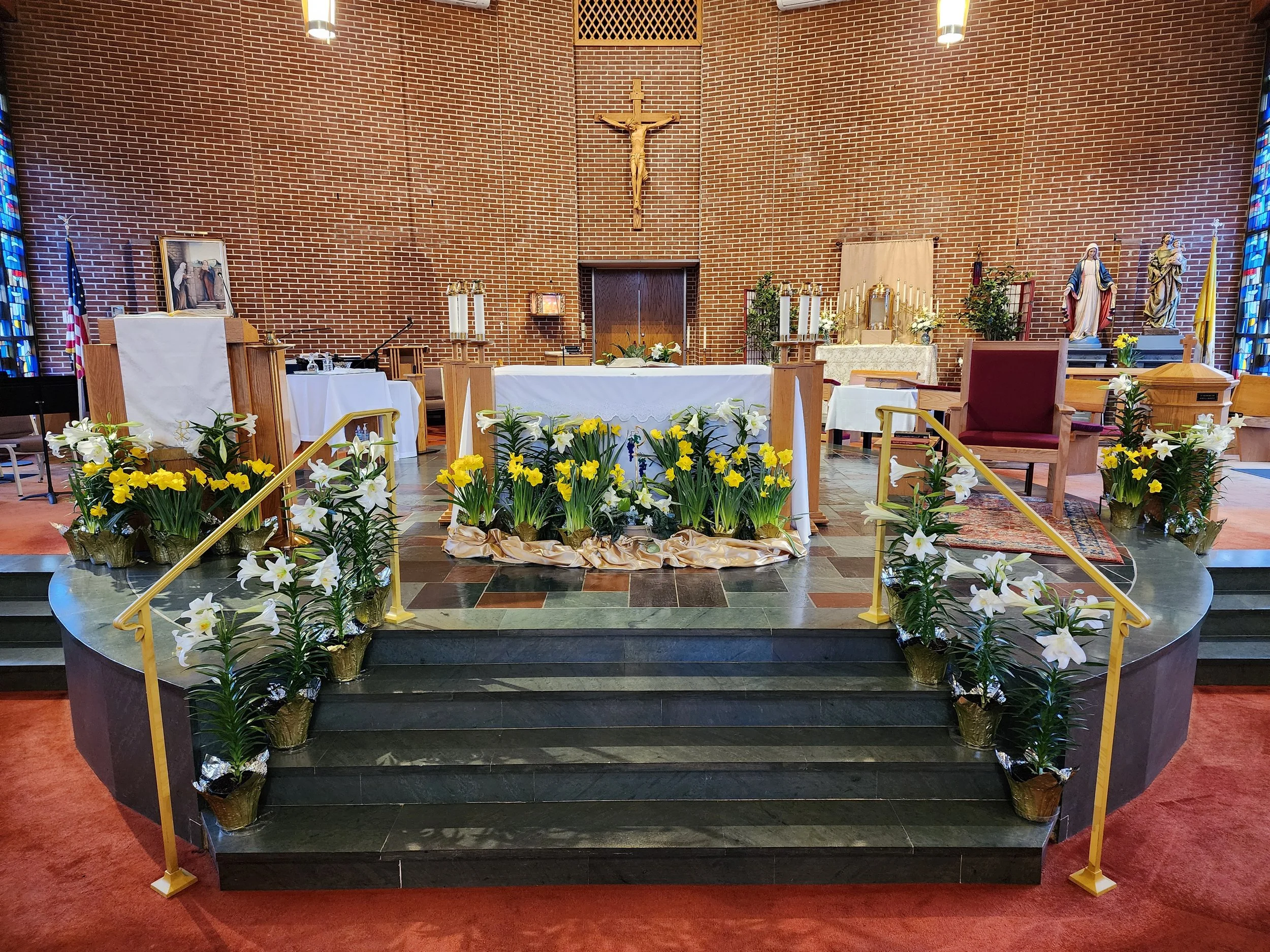Religious altar decorated with white and yellow flowers, on tiered black steps with gold-colored railings inside a church.