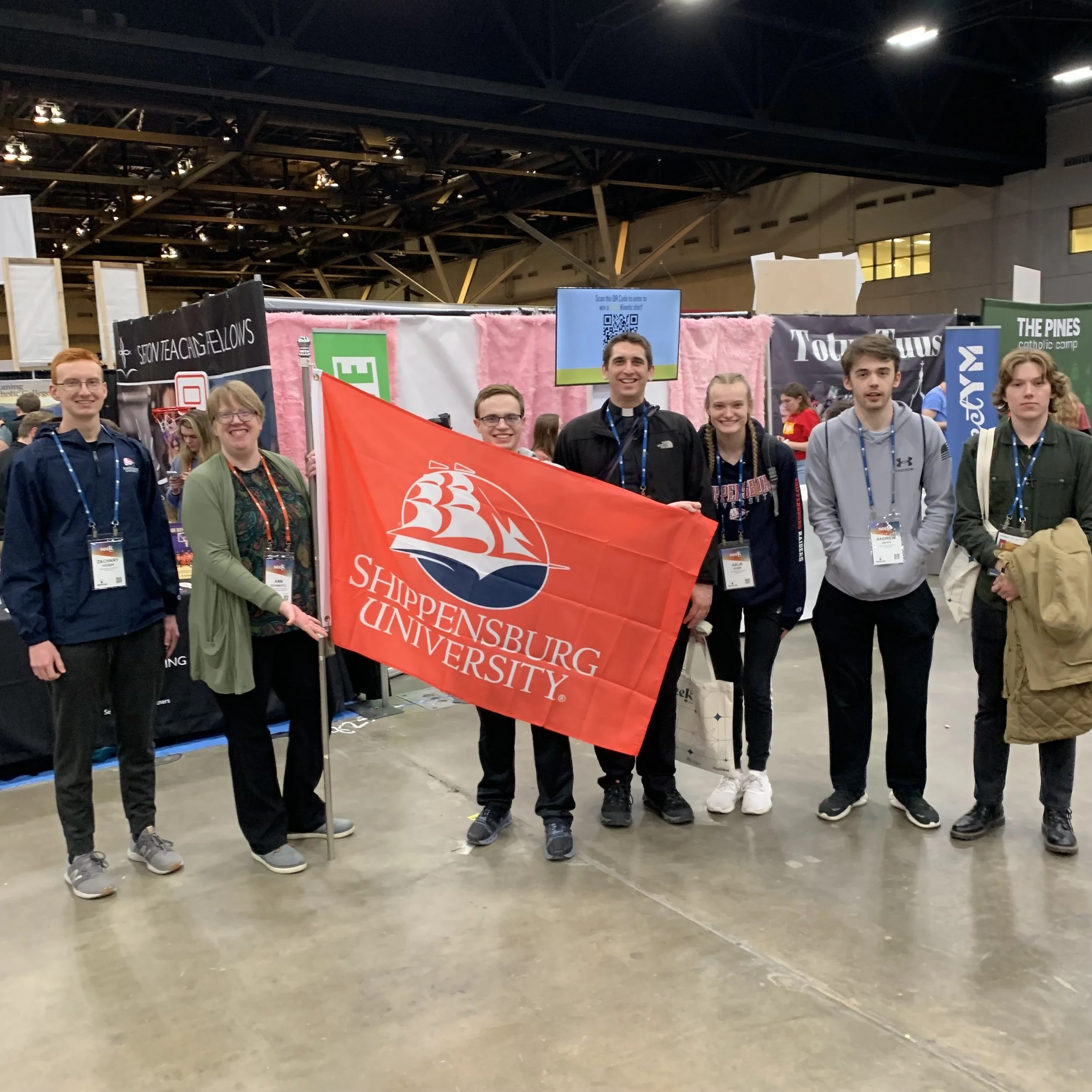 Group of seven young individuals at an indoor event, holding a Shippensburg University flag. They are standing in front of booths and banners in a spacious, industrial-style venue.