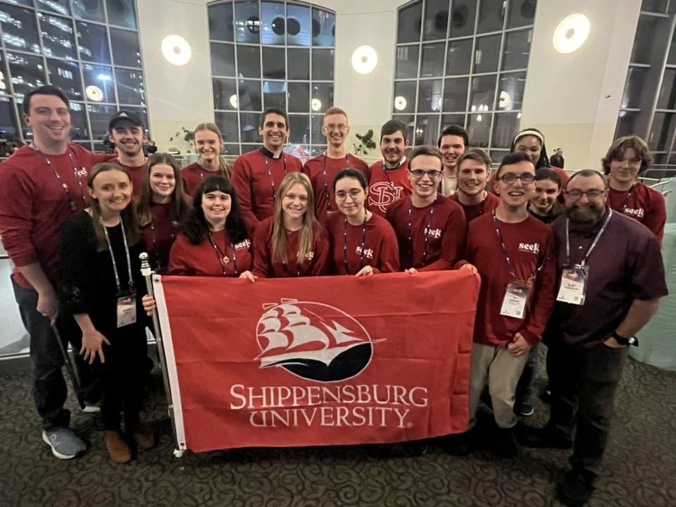 Group of young adults and a few older individuals, some wearing red jackets with 'Seek' on them, holding a red banner with the Shippensburg University logo and name, in an indoor setting with large windows and round lights.