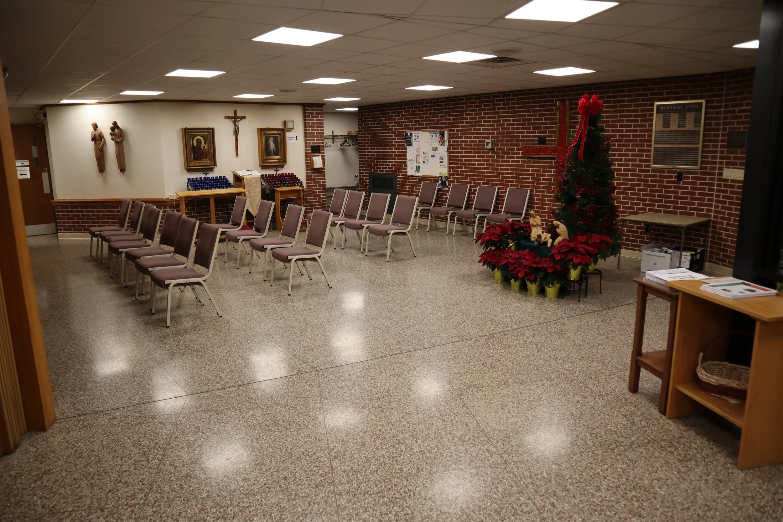 A church's basement or community room decorated for Christmas with poinsettia plants, an artificial Christmas tree with a red ribbon, religious artwork, and chairs arranged for a gathering.