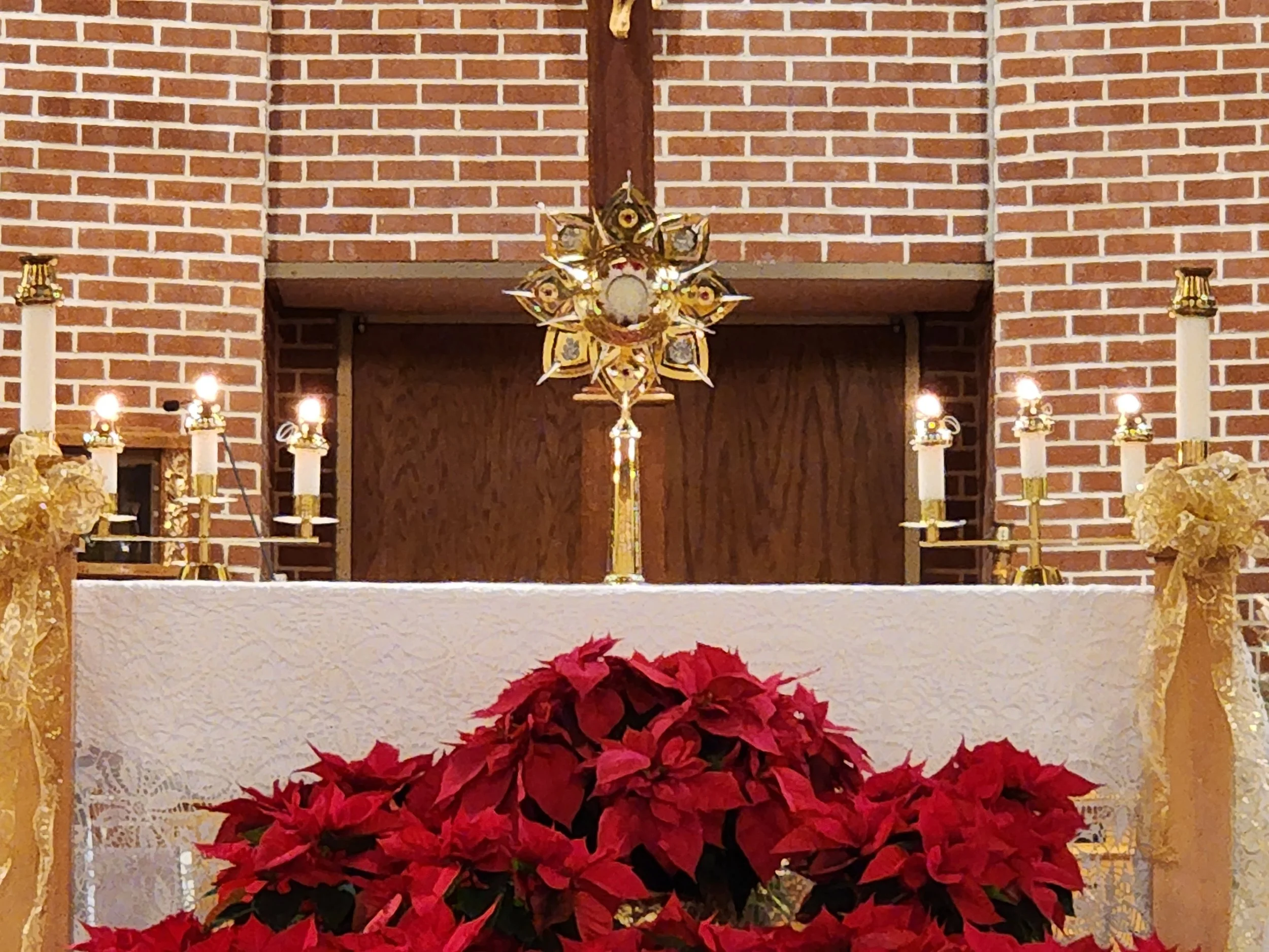 Interior of a church altar decorated with gold accents, white lace cloth, red poinsettia flowers, and lit candles, with a brick wall in the background.