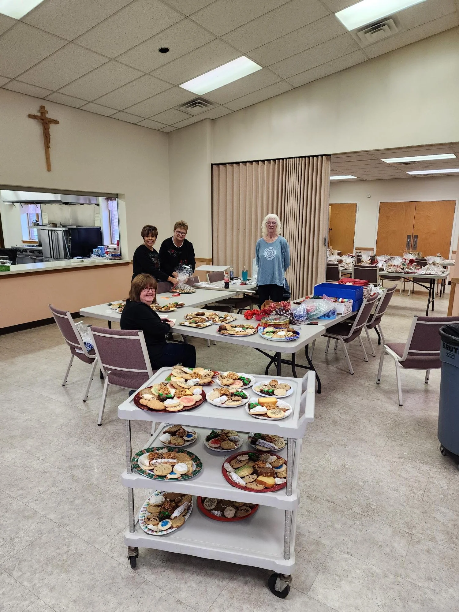 A room with four women around a table covered with plates of decorated cookies, with additional dishes on a cart in the foreground. The room has tan walls, a cross on the wall, and kitchen area in the background.