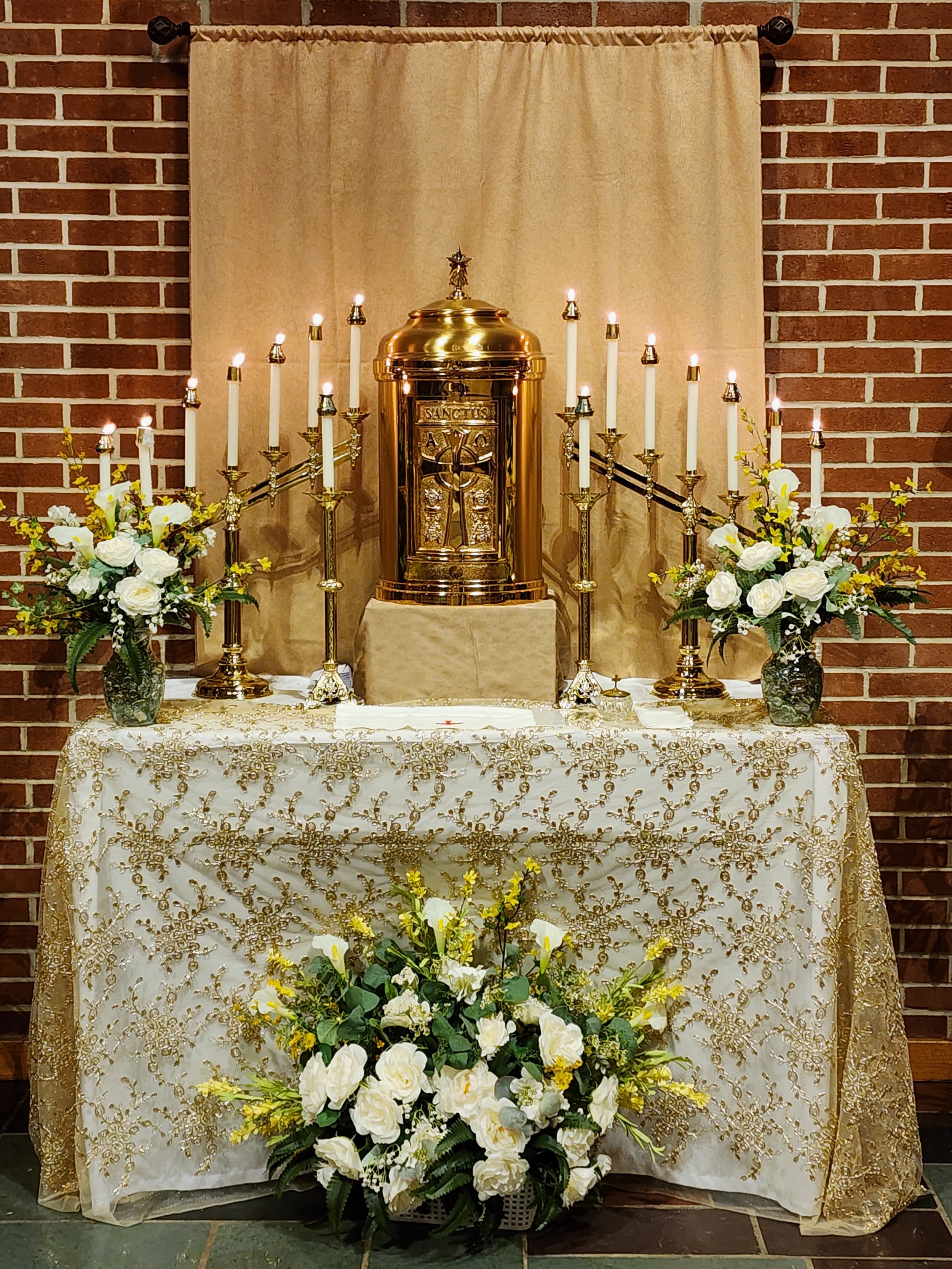 Decorated altar with a golden relic, surrounded by lit candles, flower arrangements, and a patterned white and gold cloth, set against a brick wall with beige backdrop.
