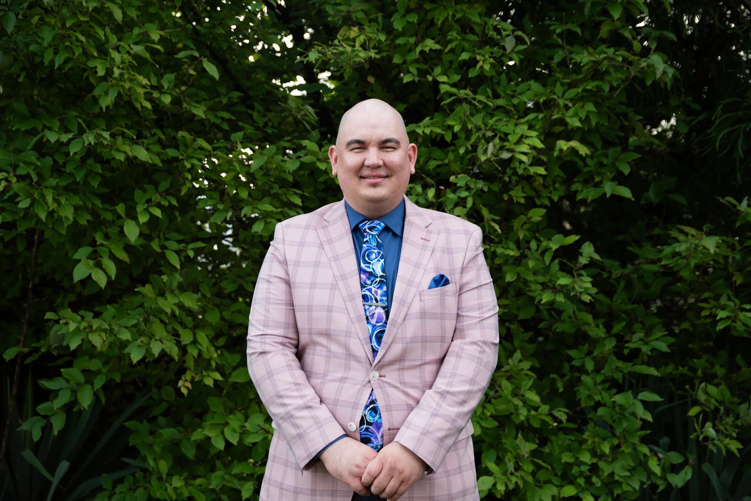 A bald man in a pink plaid suit with a blue dress shirt, a blue patterned tie, and a blue pocket square standing in front of green foliage, smiling at the camera.