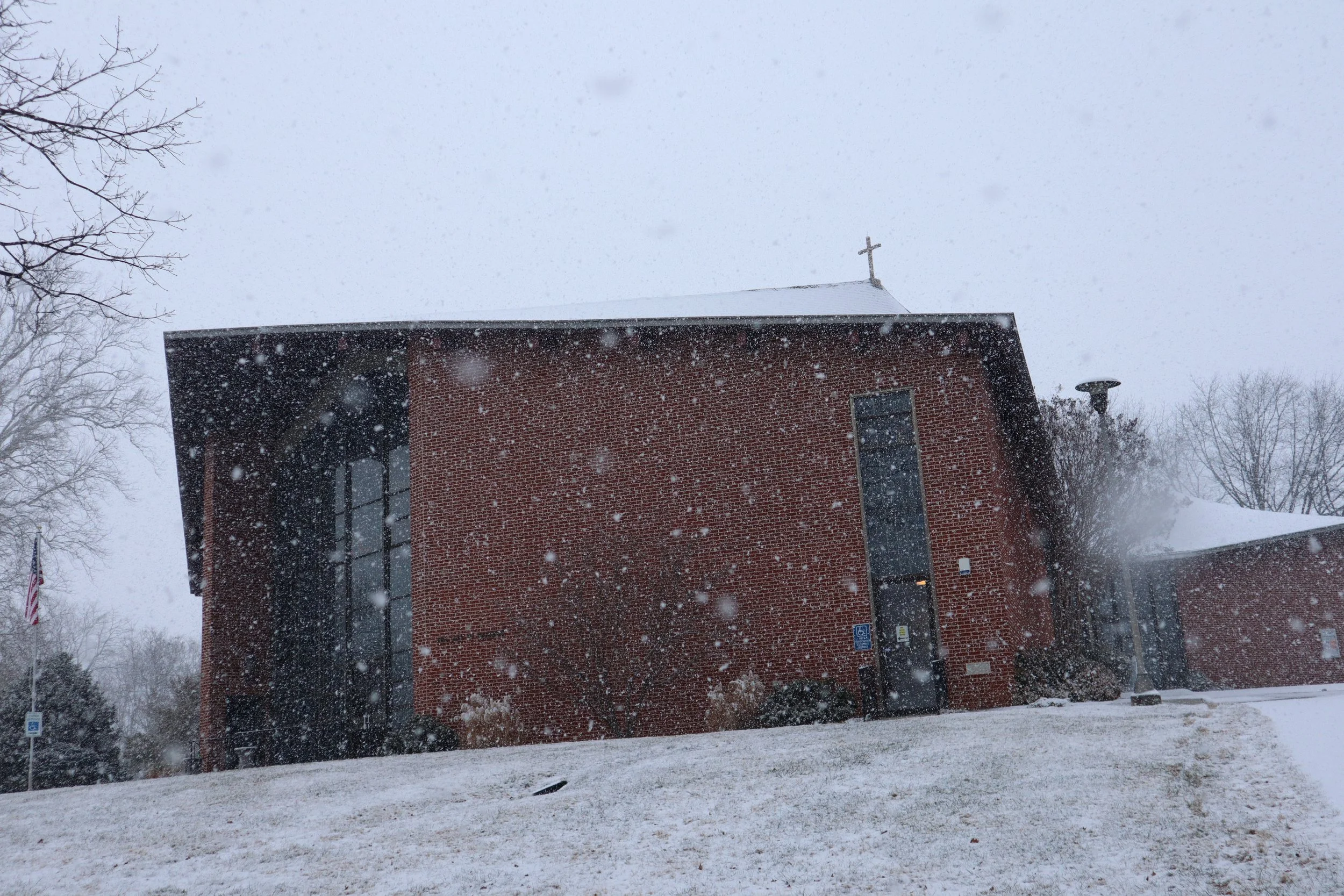 Brick church building in a snowy landscape with a cross on top and snow falling.
