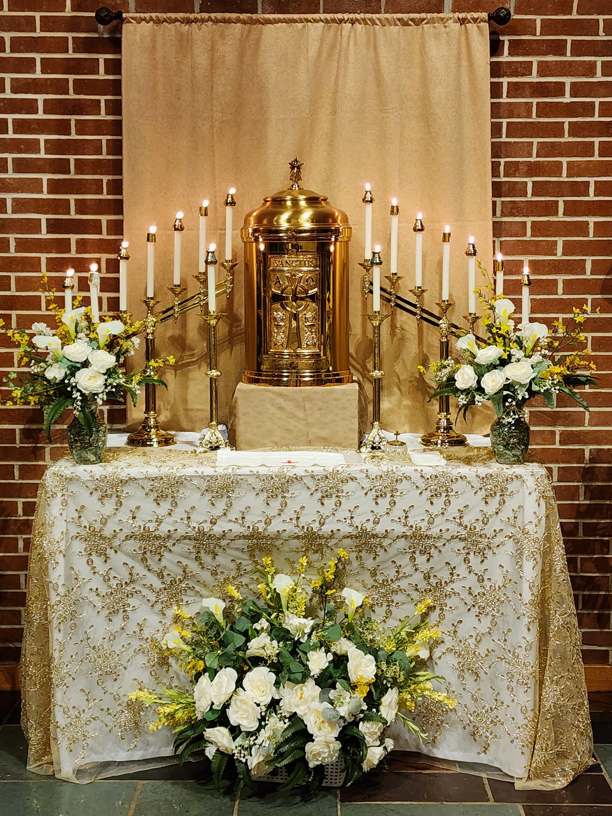 An altar decorated with white and gold, featuring a gold tabernacle in the center, surrounded by 16 lit candles on gold holders and white floral arrangements on either side and at the base, against a brick wall with a beige curtain backdrop.