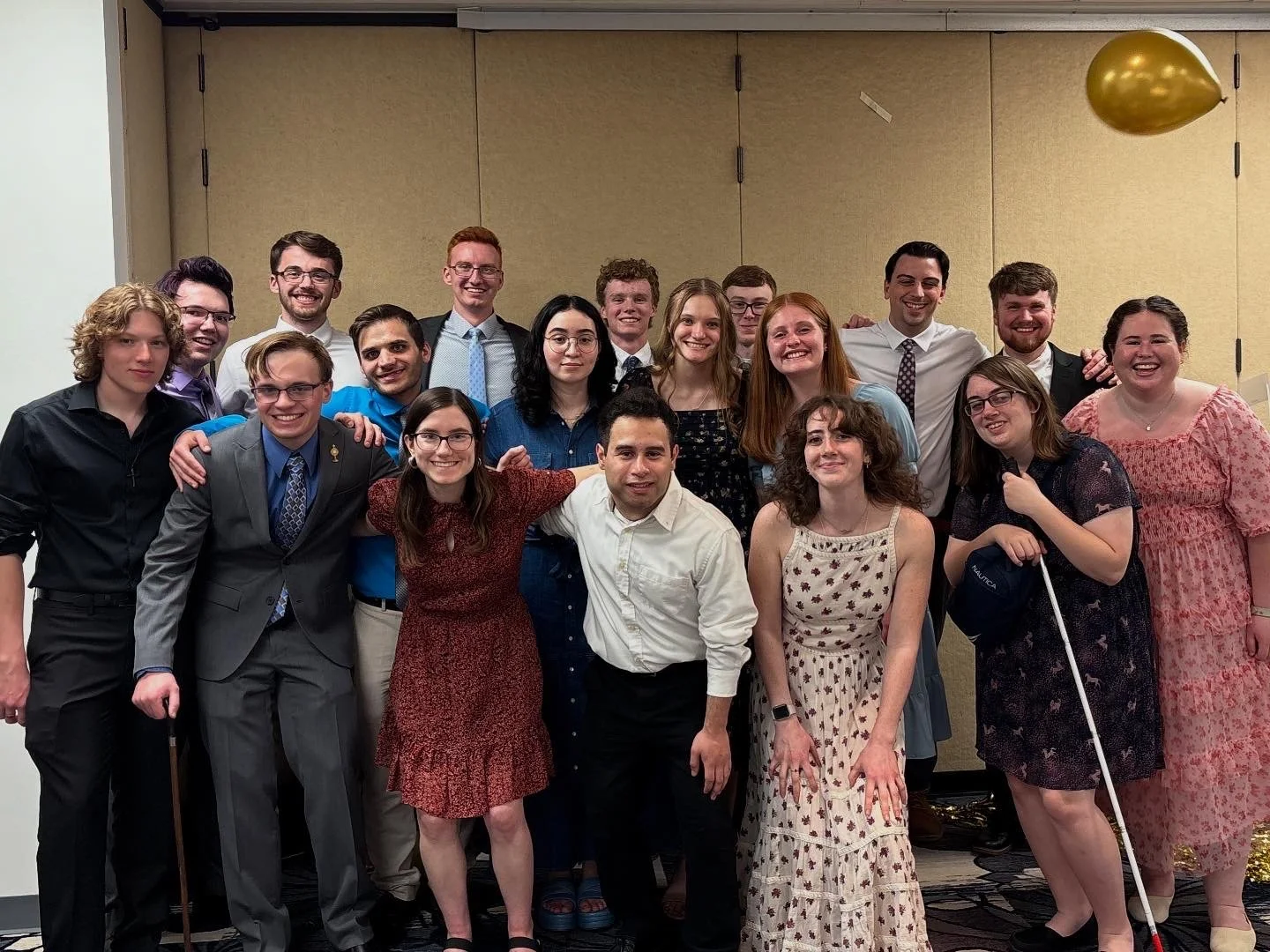 Group of 17 young adults in formal and casual attire, smiling and posing together at an indoor event with beige walls and a gold balloon in the background.