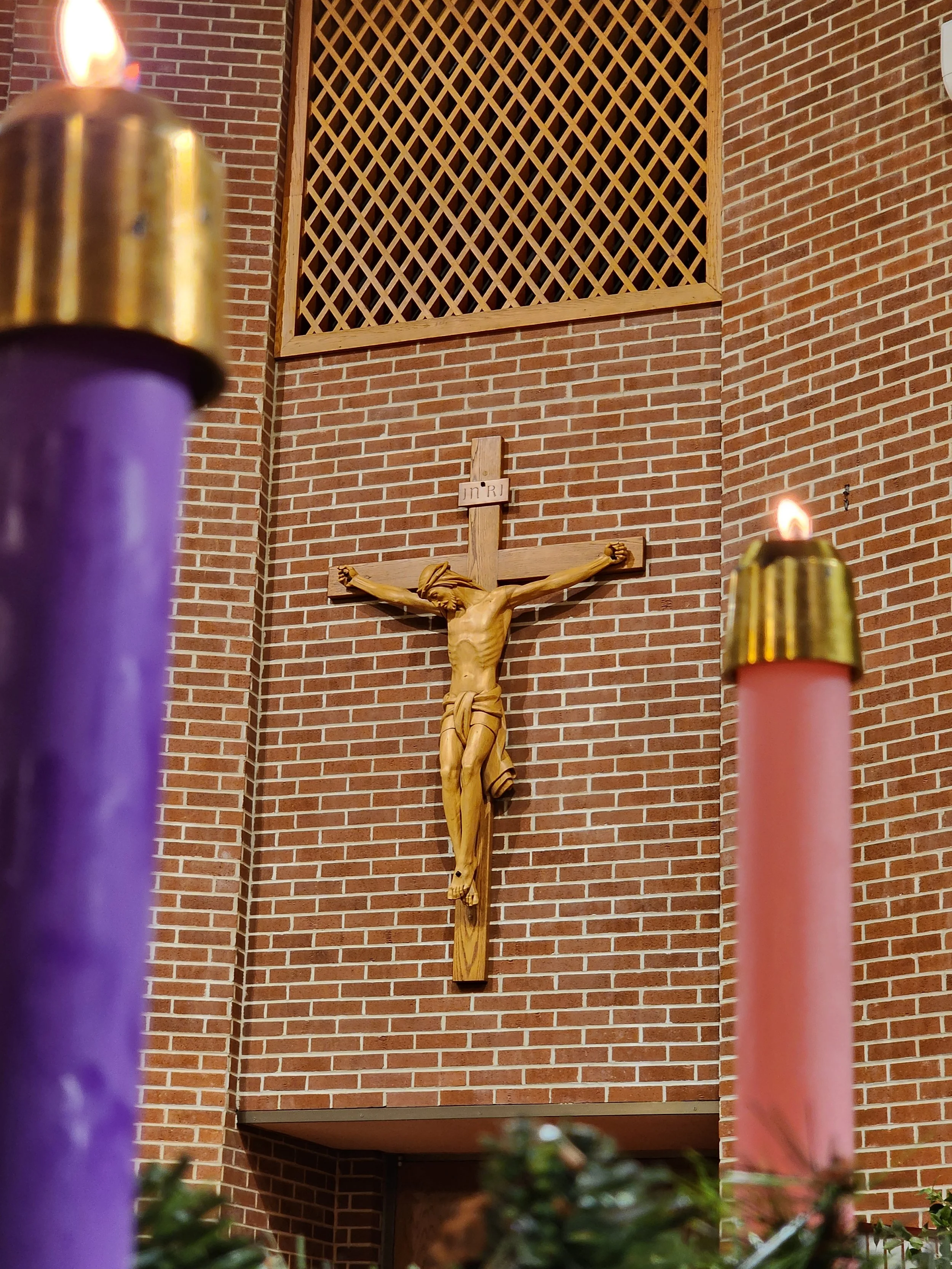 A wooden crucifix with Jesus Christ on it mounted on a brick wall inside a church, with two lit candles in purple and pink holders in the foreground.
