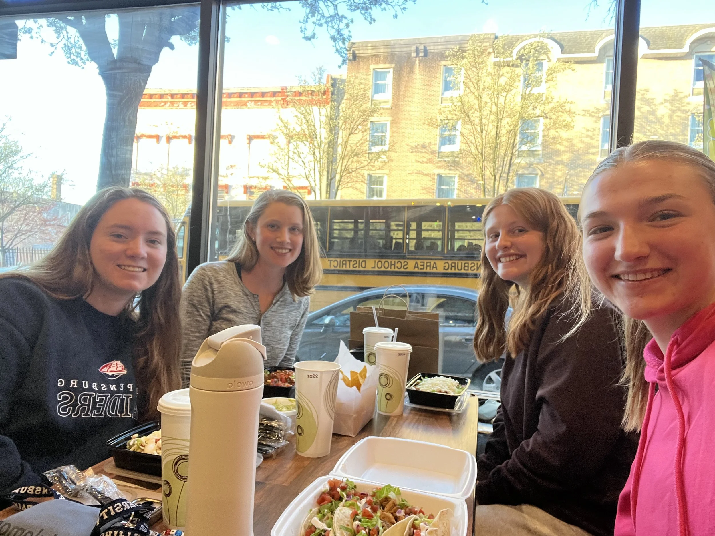 Four young girls sitting at a dining table inside a restaurant, smiling at the camera with food and drinks in front of them. Outside through the window, a yellow school bus and trees are visible.