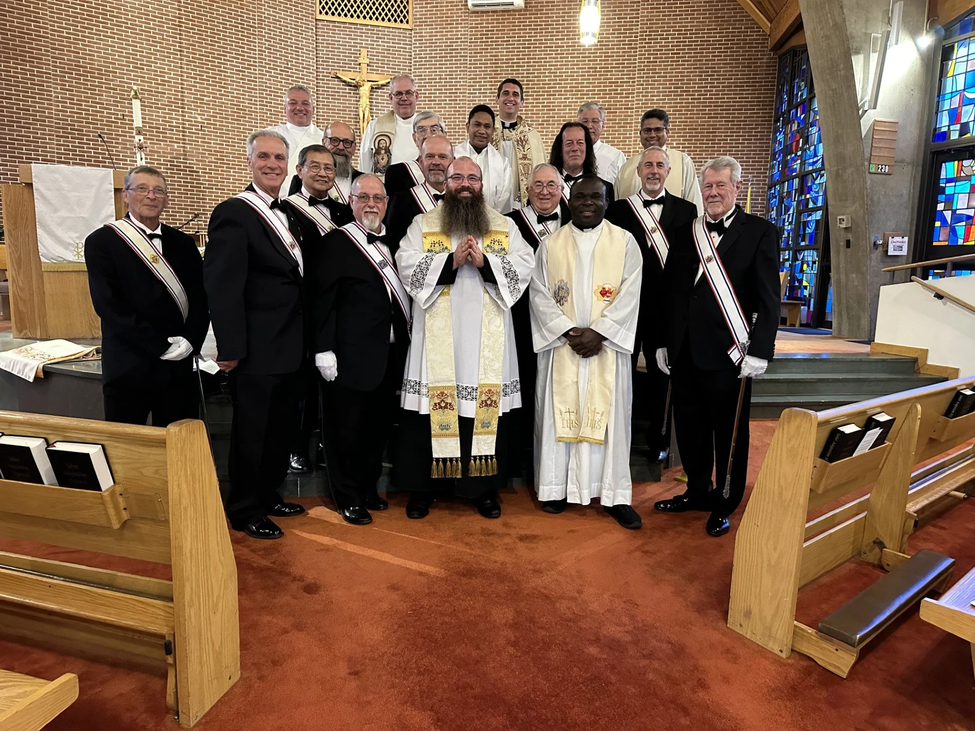Group of clergy and church officials posing inside a church with stained glass windows and a crucifix in the background.