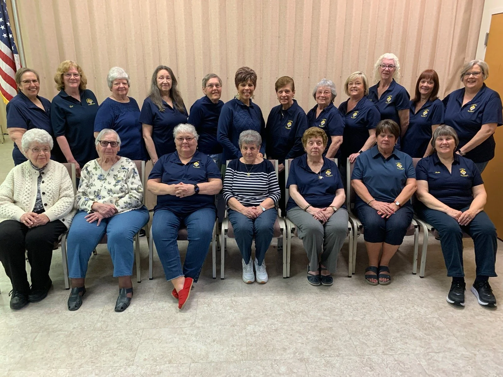 Group of 16 women, some sitting and some standing, posing indoors in front of a beige curtain. Most are wearing navy blue shirts with a logo, with a few in casual or different attire. An American flag is partially visible on the far left.