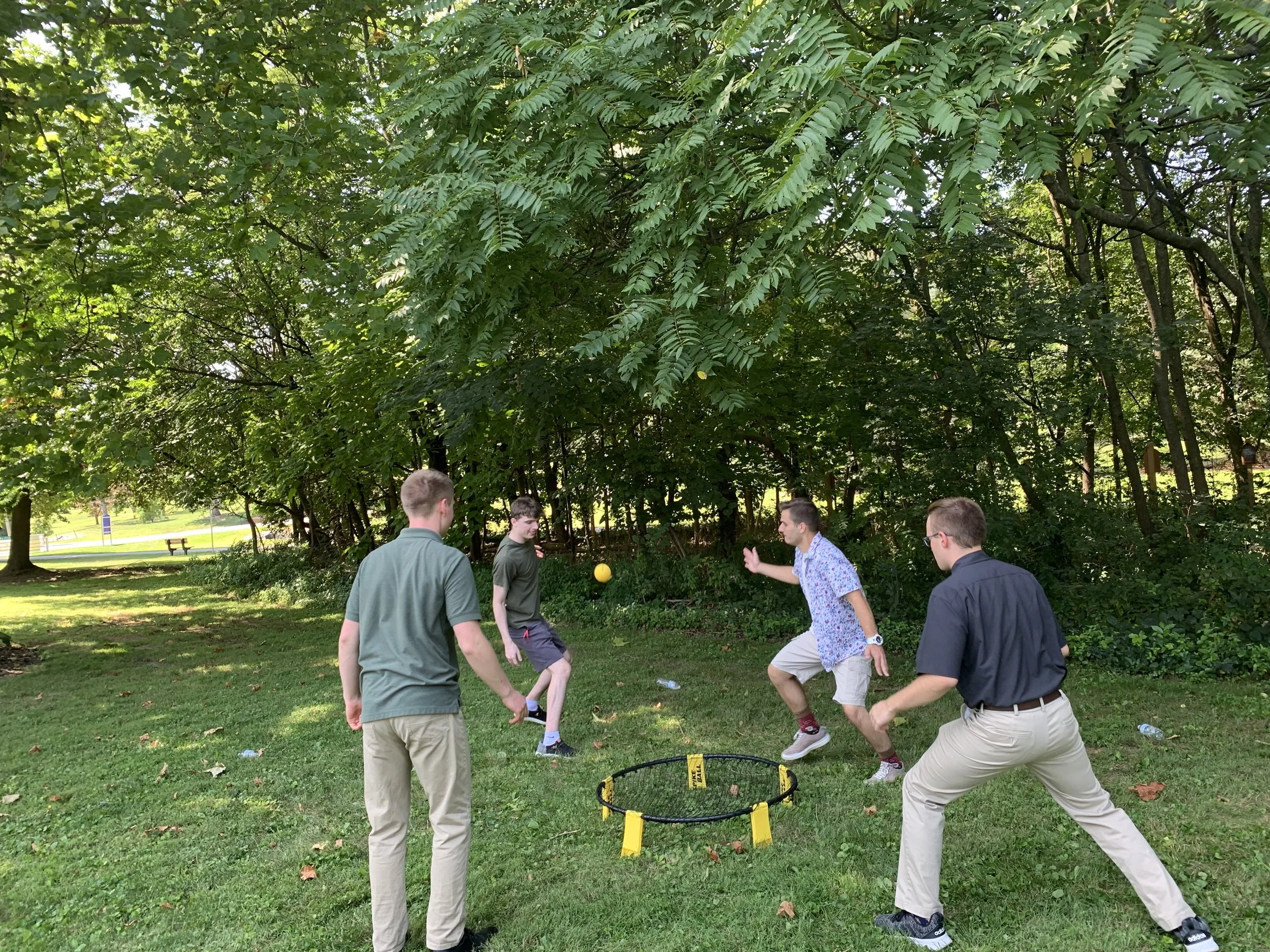 Four young men playing cornhole outdoors on a grassy area near trees, with a yellow bean bag in mid-air and a yellow and black cornhole game set on the ground.