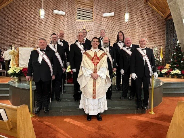A group of men dressed in formal attire and sashes, standing on the steps of a church altar with a priest in liturgical vestments, inside a church decorated with flowers and Christmas trees.