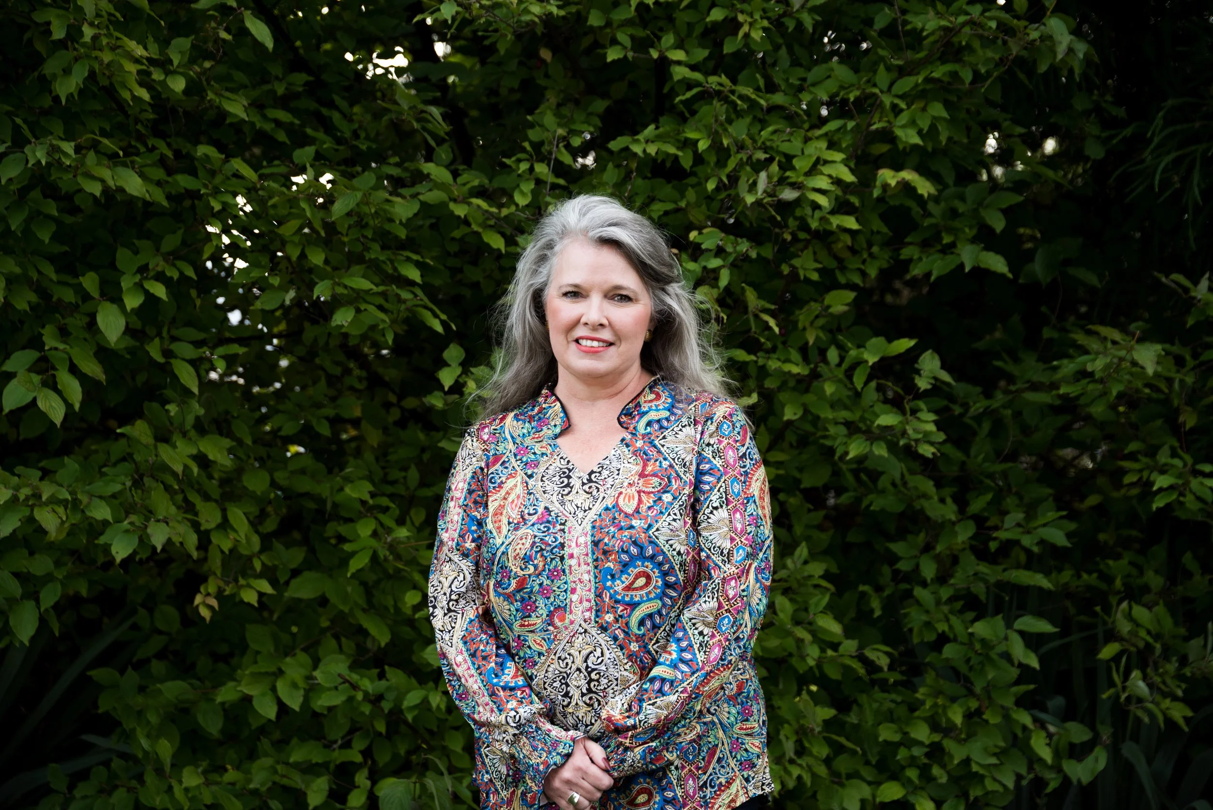A woman with long gray hair standing outdoors in front of green leafy bushes, wearing a colorful patterned blouse.