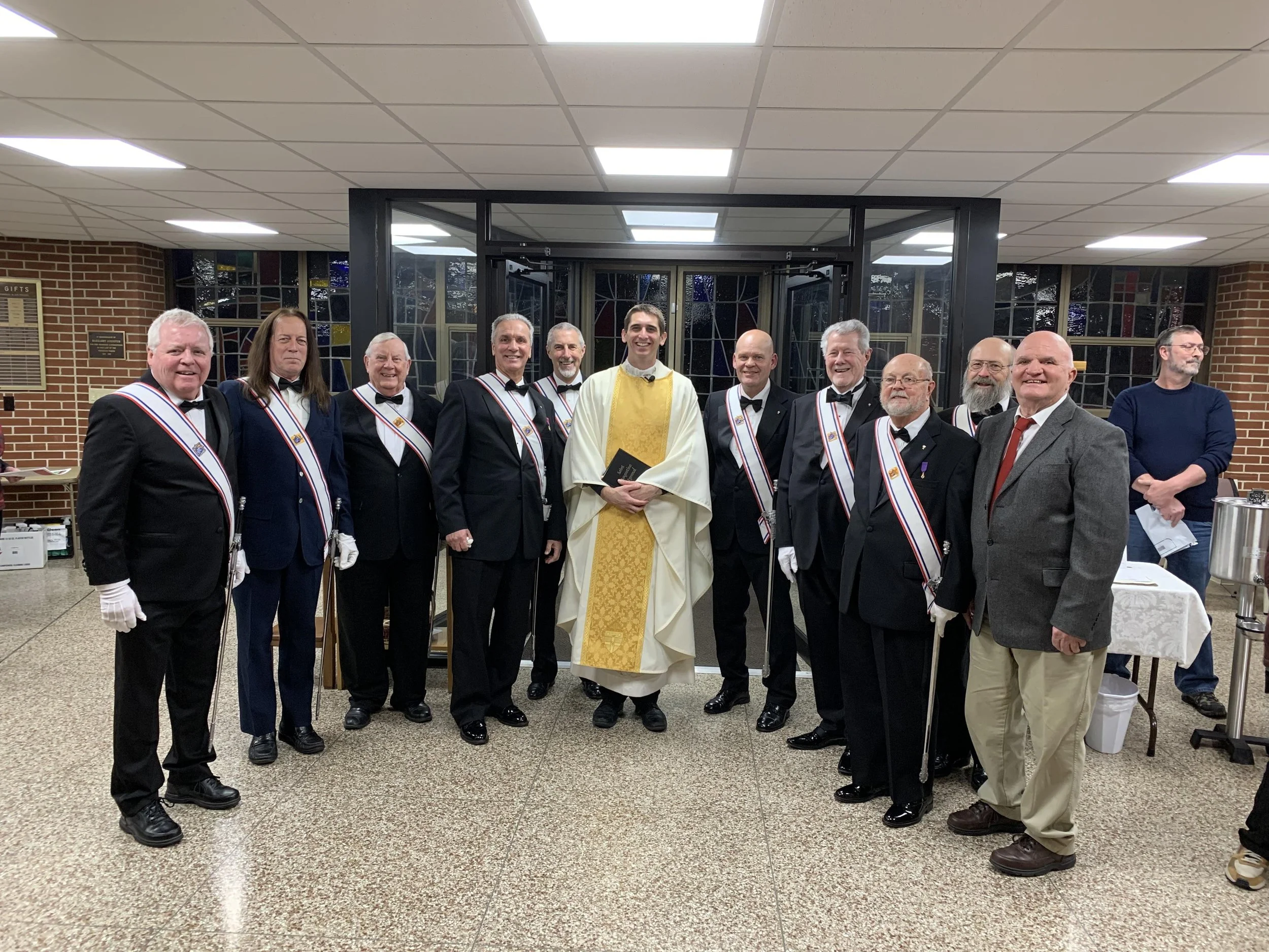A group of men and one person in priestly robes, all dressed in formal attire, standing indoors in front of glass doors, celebrating an event. Some men are wearing sashes and gloves, and everyone appears to be smiling.