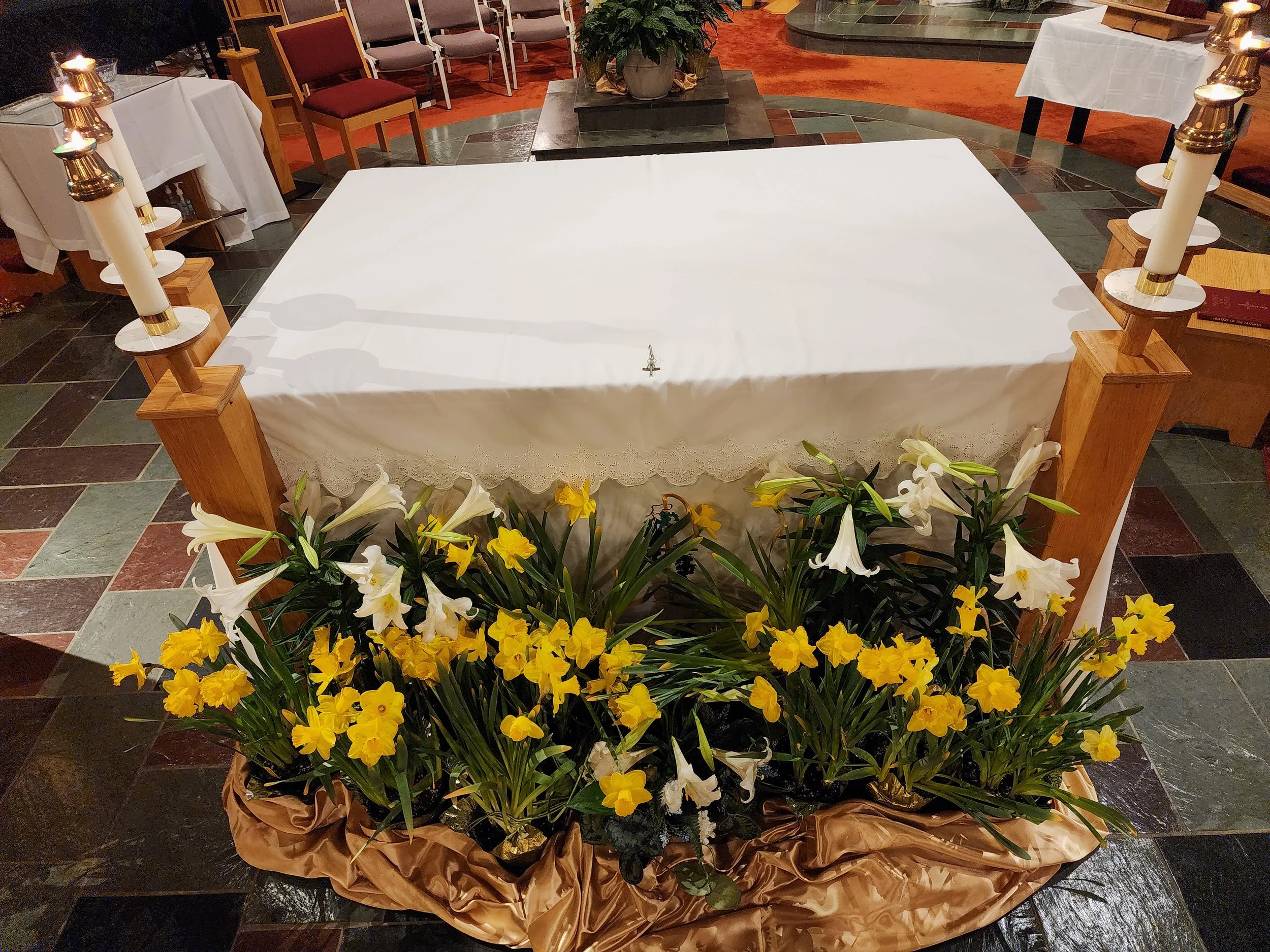 A religious altar with a white cloth, surrounded by yellow and white flowers, on a tiled floor with chairs and candles nearby.