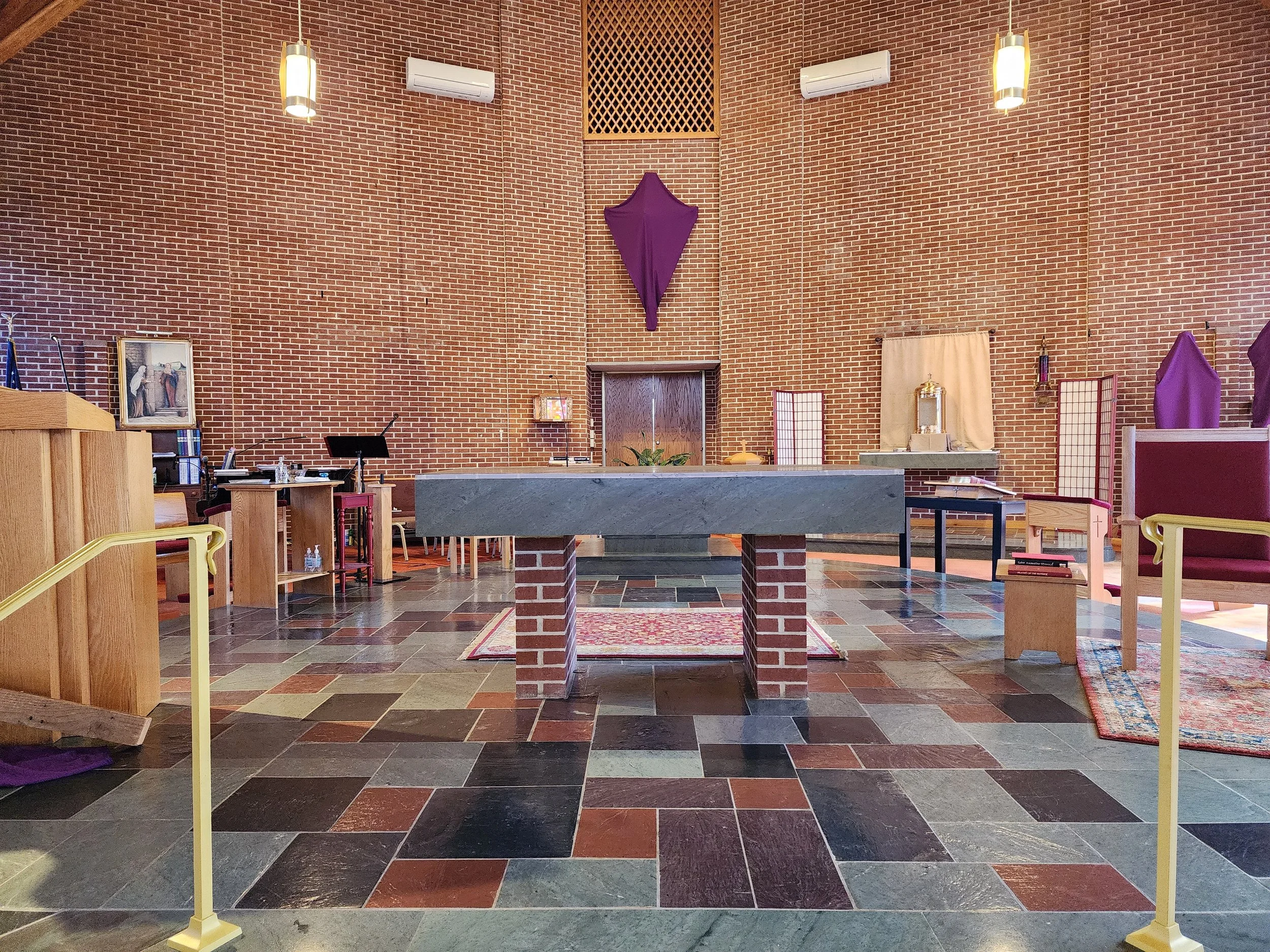 Interior of a church with a brick wall, a central altar with a gray top, purple drapes, wooden chairs, and religious items. The floor is tiled with various colored stones.