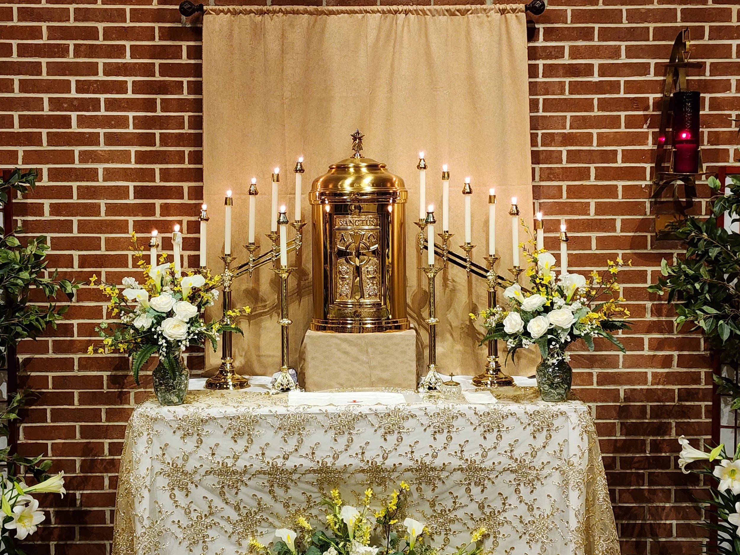 An altar decorated with white and yellow flowers, multiple lit candles, and a golden tabernacle against a beige backdrop on a table with gold embroidery, set against a brick wall.