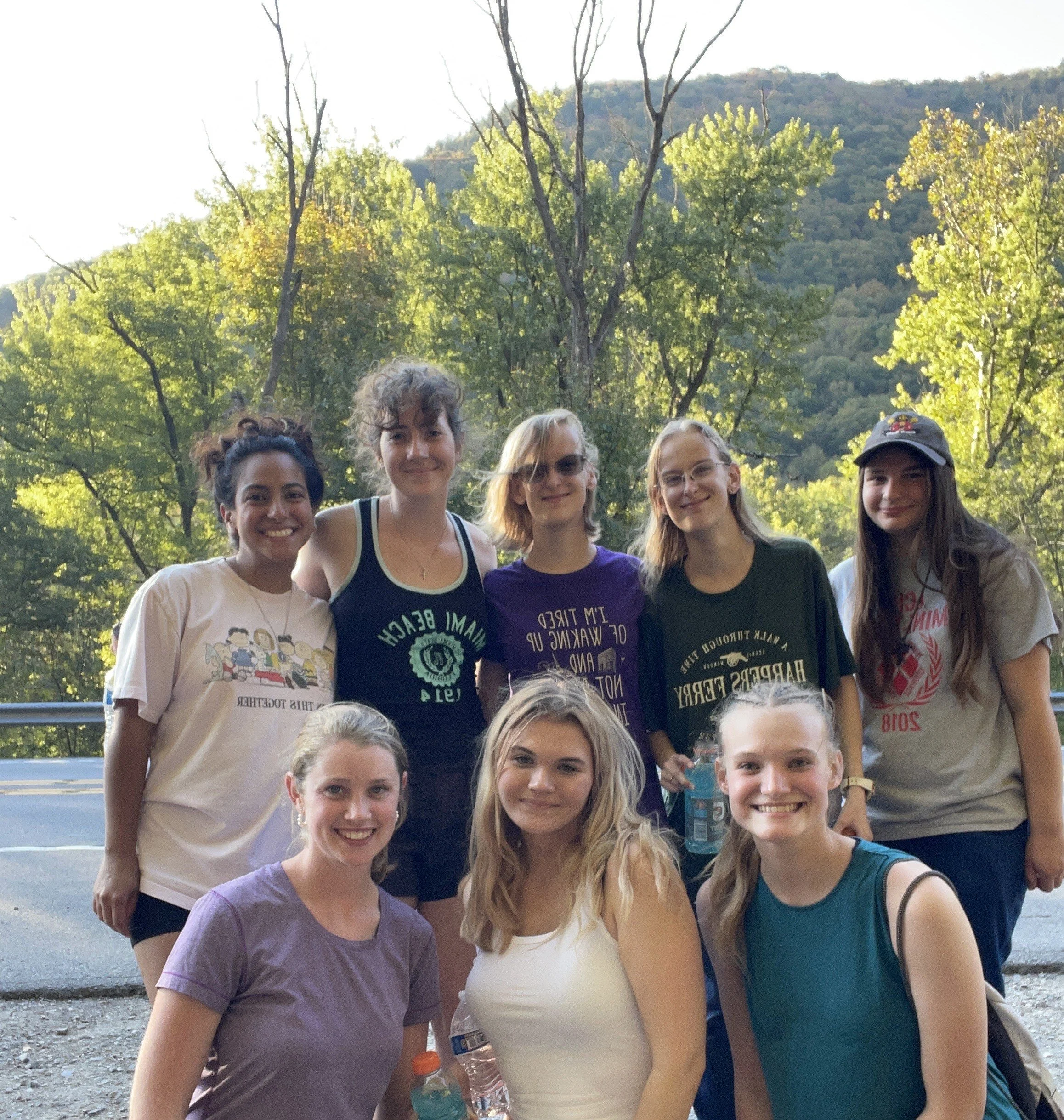 Group of ten young women smiling outdoor in a forested area with mountains in the background.