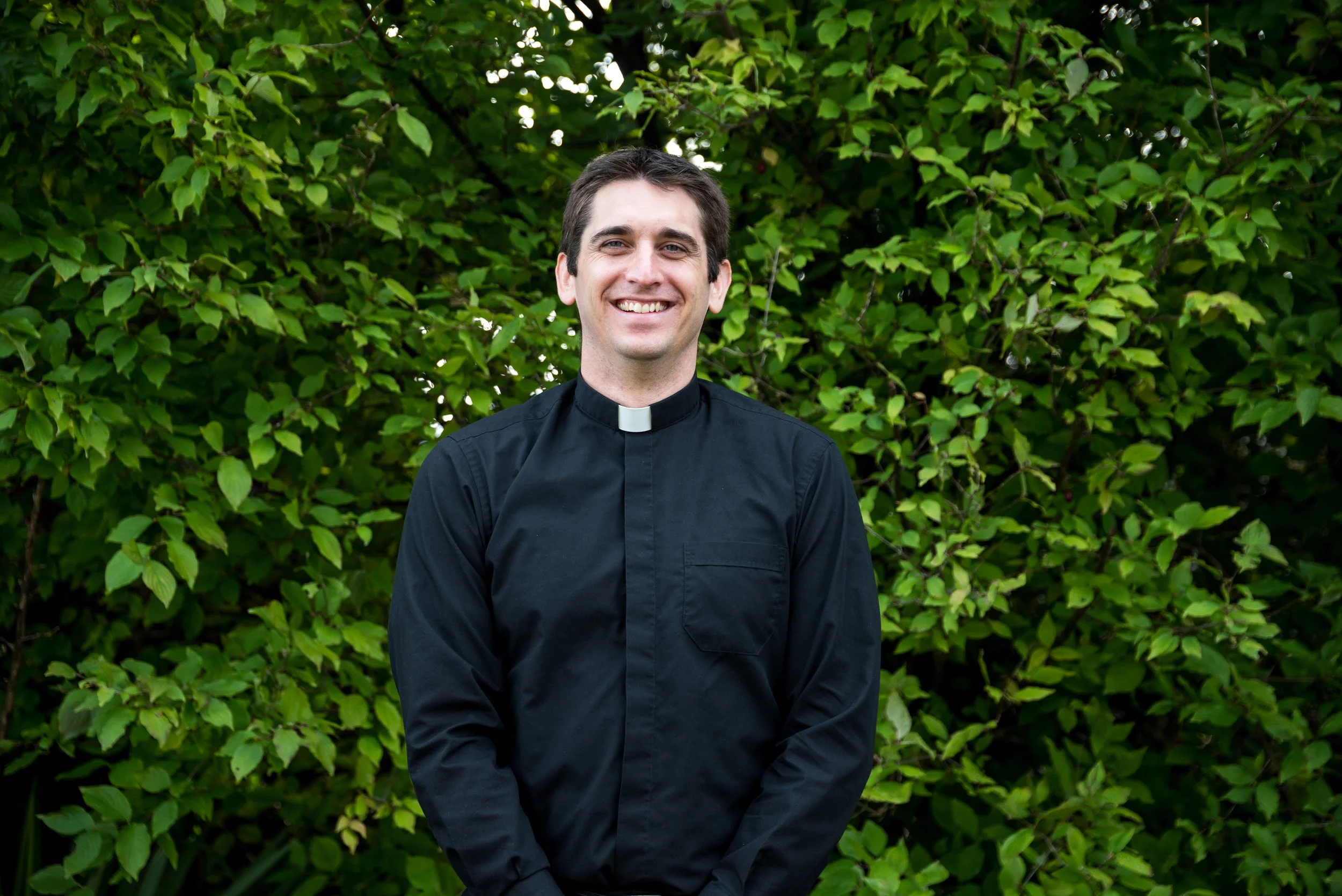 A smiling man wearing a black clergy shirt with a white clerical collar standing outdoors in front of green foliage.