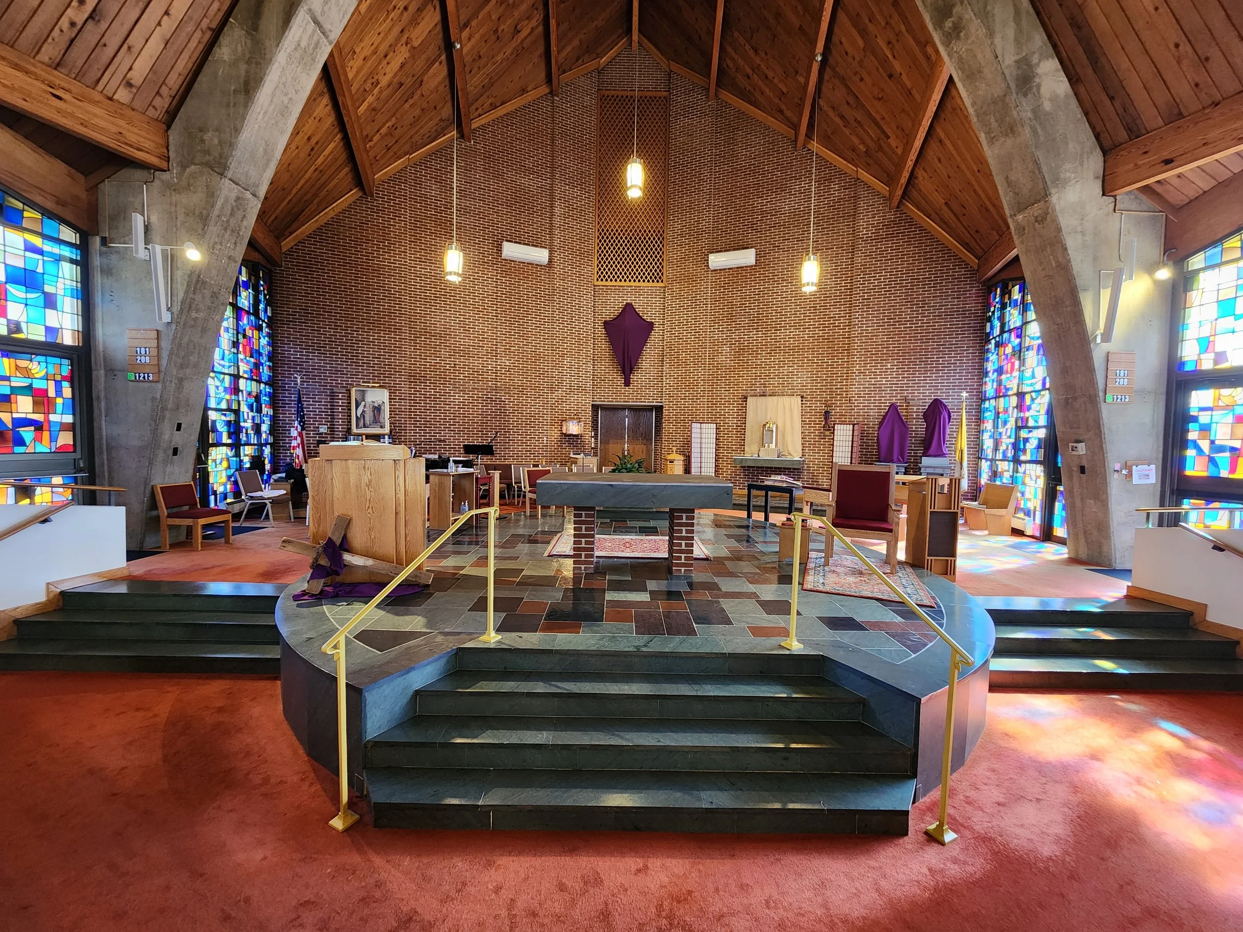 Interior of a church altar with a brick wall, stained glass windows, wooden ceiling, and purple altar cloths. The altar is on a raised platform with steps, and there are chairs and chairs with purple coverings around.