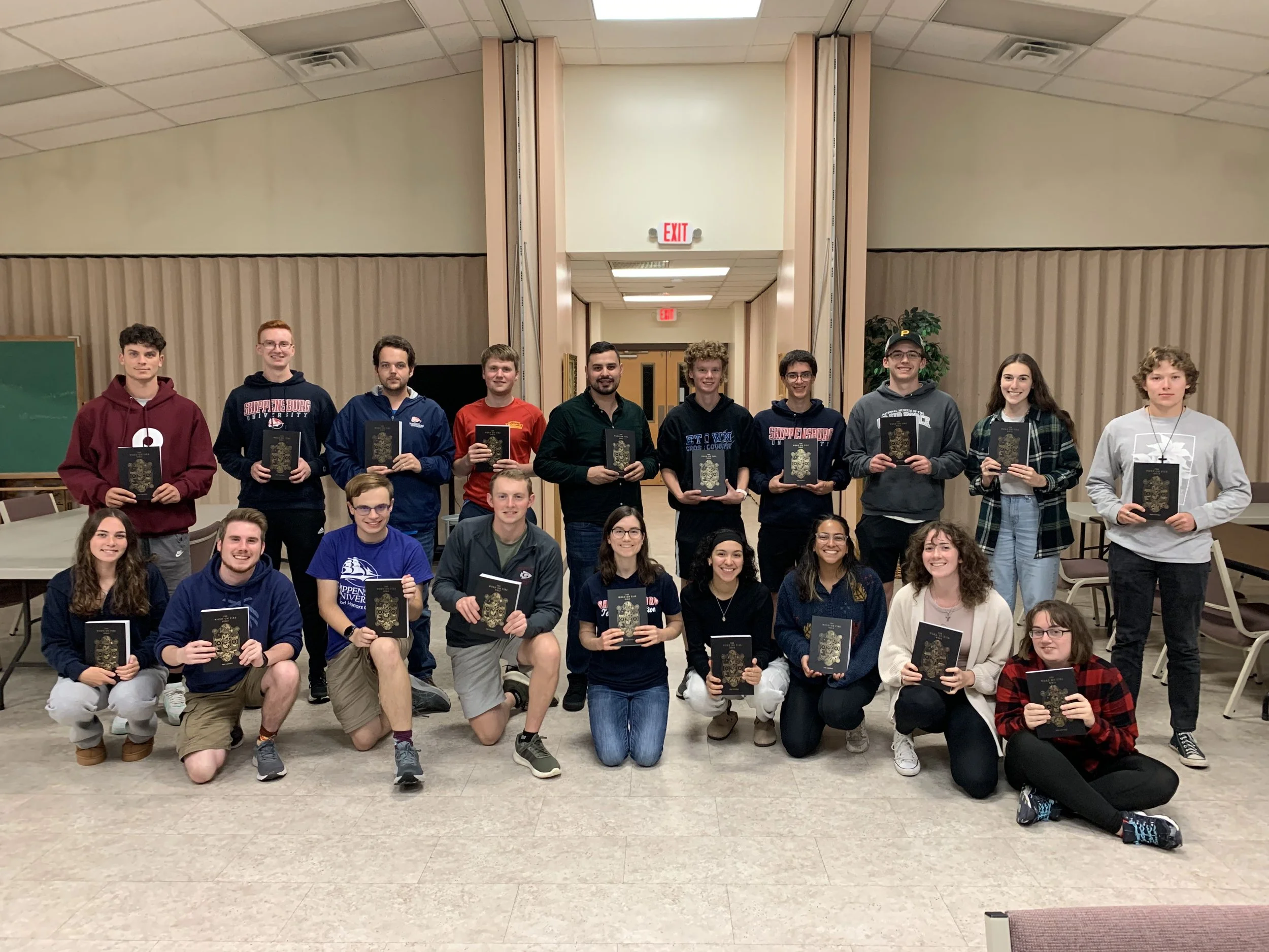Group of twenty-one young adults, standing and kneeling indoors in a room with beige walls, holding awards or certificates, smiling for a group photo.