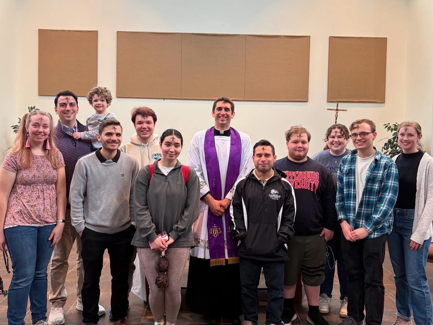 A group of fifteen people, including a priest in white robes with a purple stole, standing inside a church, smiling for a photo. The group includes men, women, and children of diverse appearances, with some holding hands or resting their hands in fro