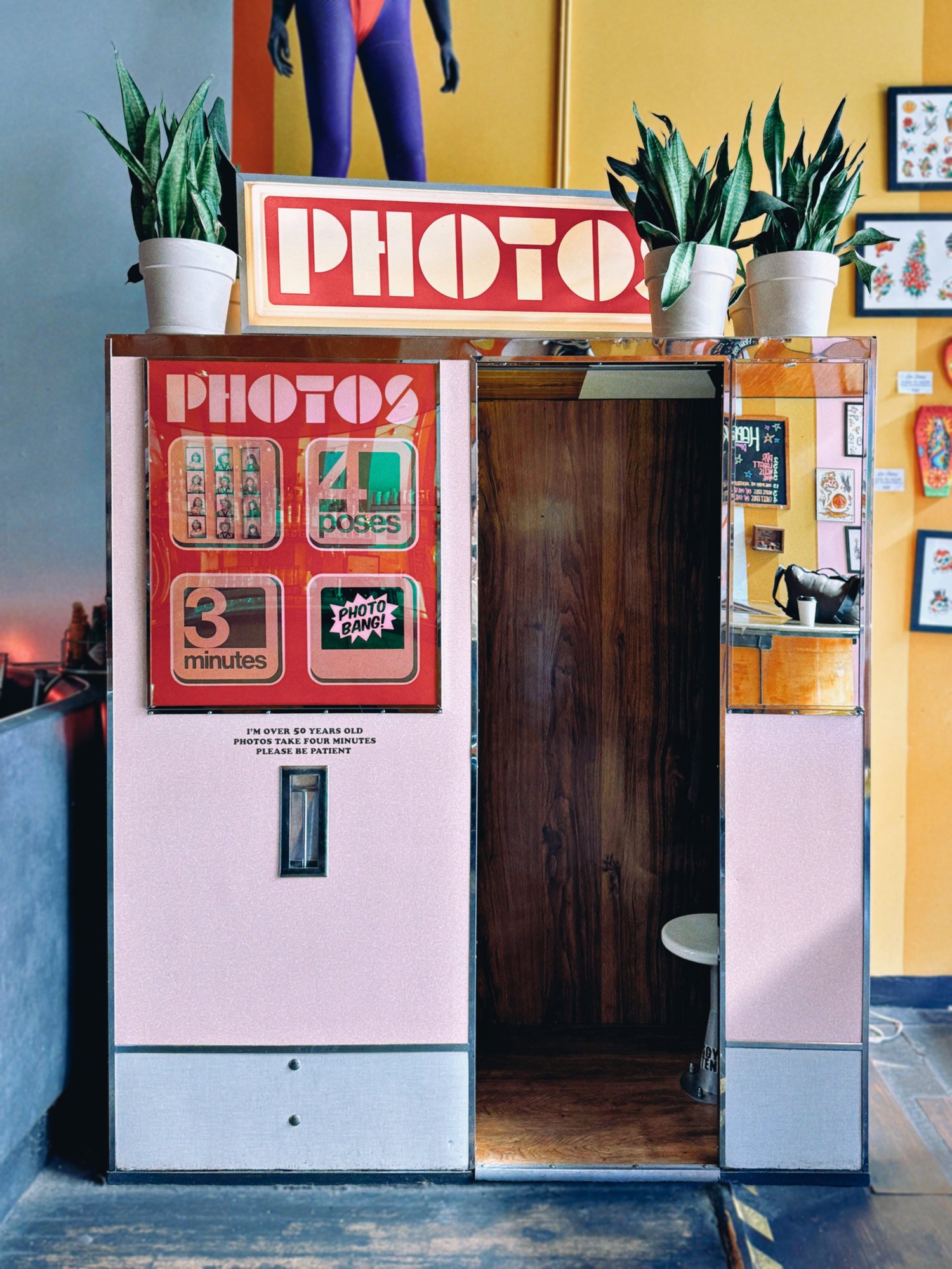 Auto-Photo Model 17 photo booth with vintage sign that says 'PHOTOS' in bold letters, decorated with potted plants, located inside a colorful room with framed pictures on the walls.