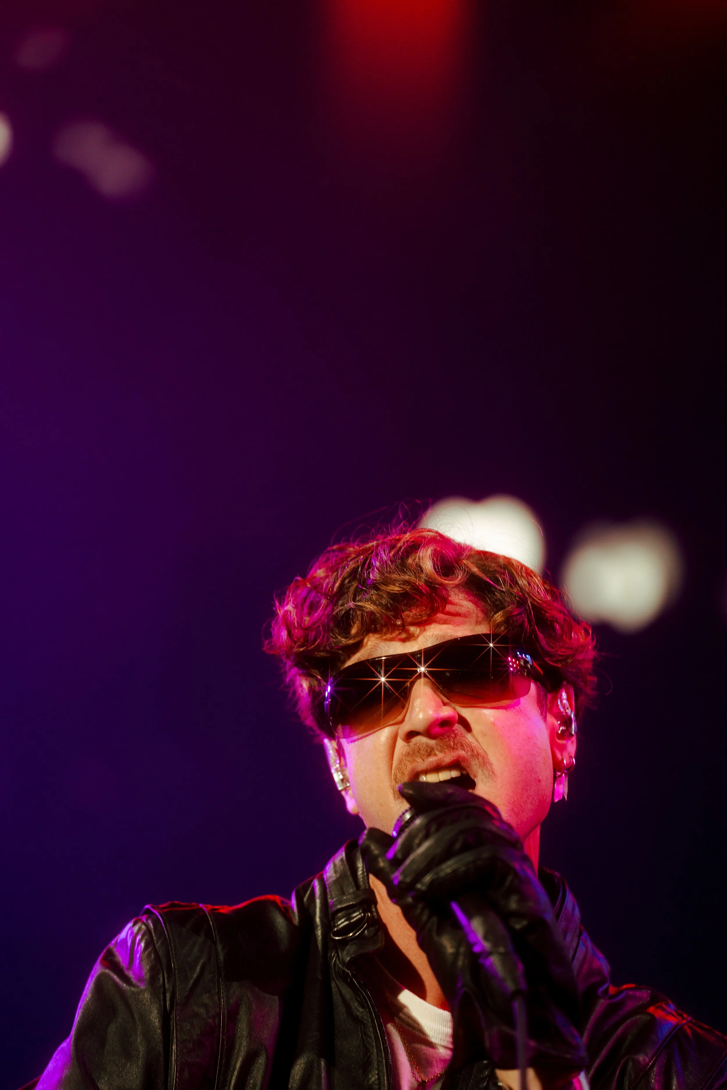 A performer singing into a microphone on stage, wearing dark sunglasses, earrings, a leather jacket, and black gloves, with colorful stage lights behind him.