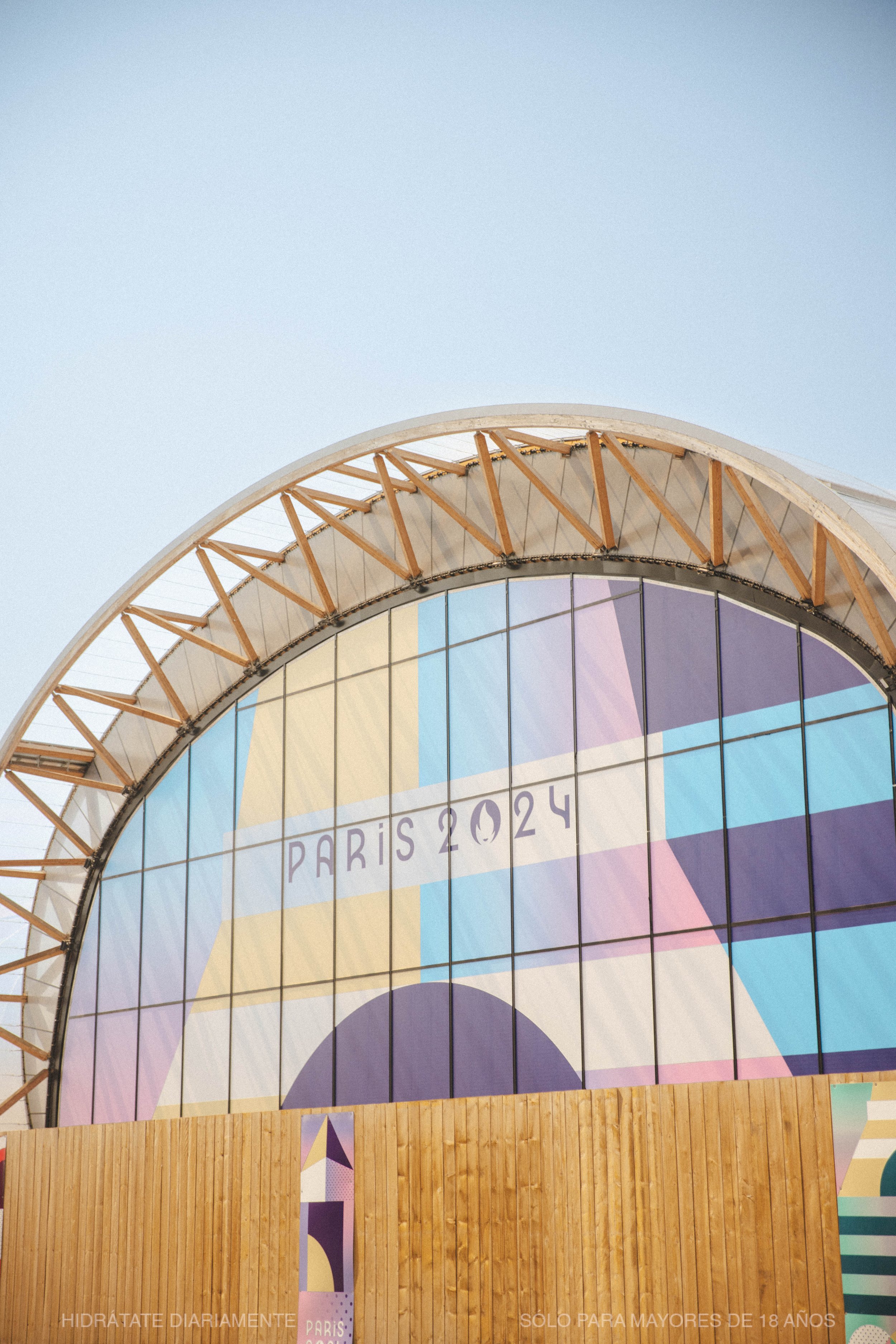 Close-up photo of a colorful building with a wooden lower section and a glass upper section displaying 'Paris 2024' in the window, indicating a venue for the 2024 Paris Olympics, against a clear sky.