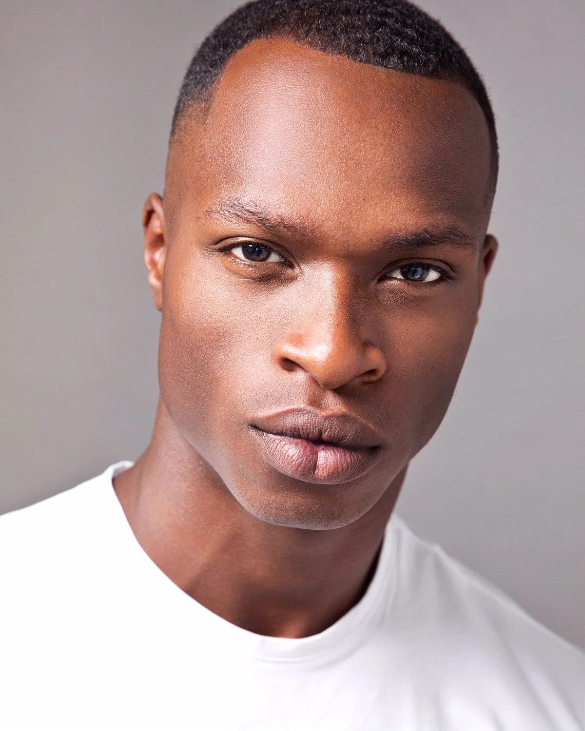 Close-up portrait of a young black man with short hair, wearing a white shirt, against a neutral gray background.
