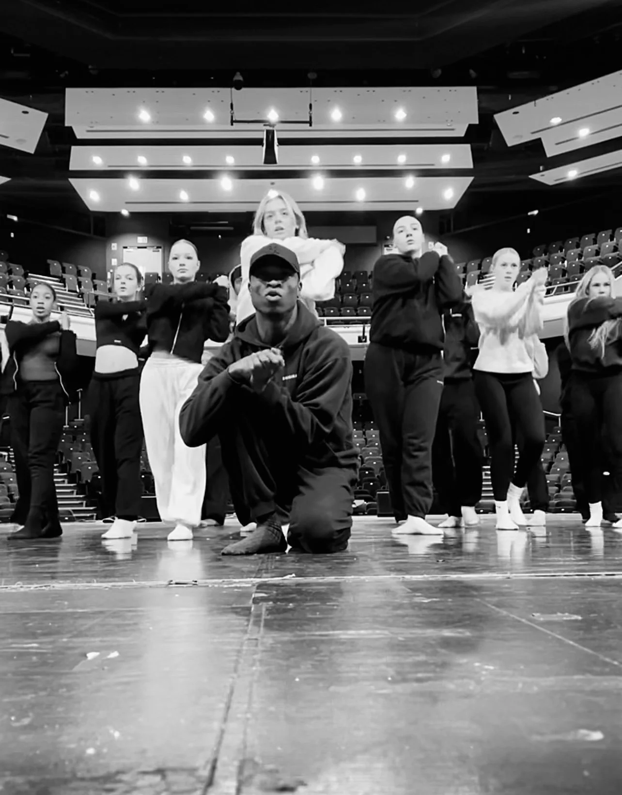 Group of dancers practicing on a stage with a person kneeling in front. The dancers are in various positions, and the setting appears to be a rehearsal space.