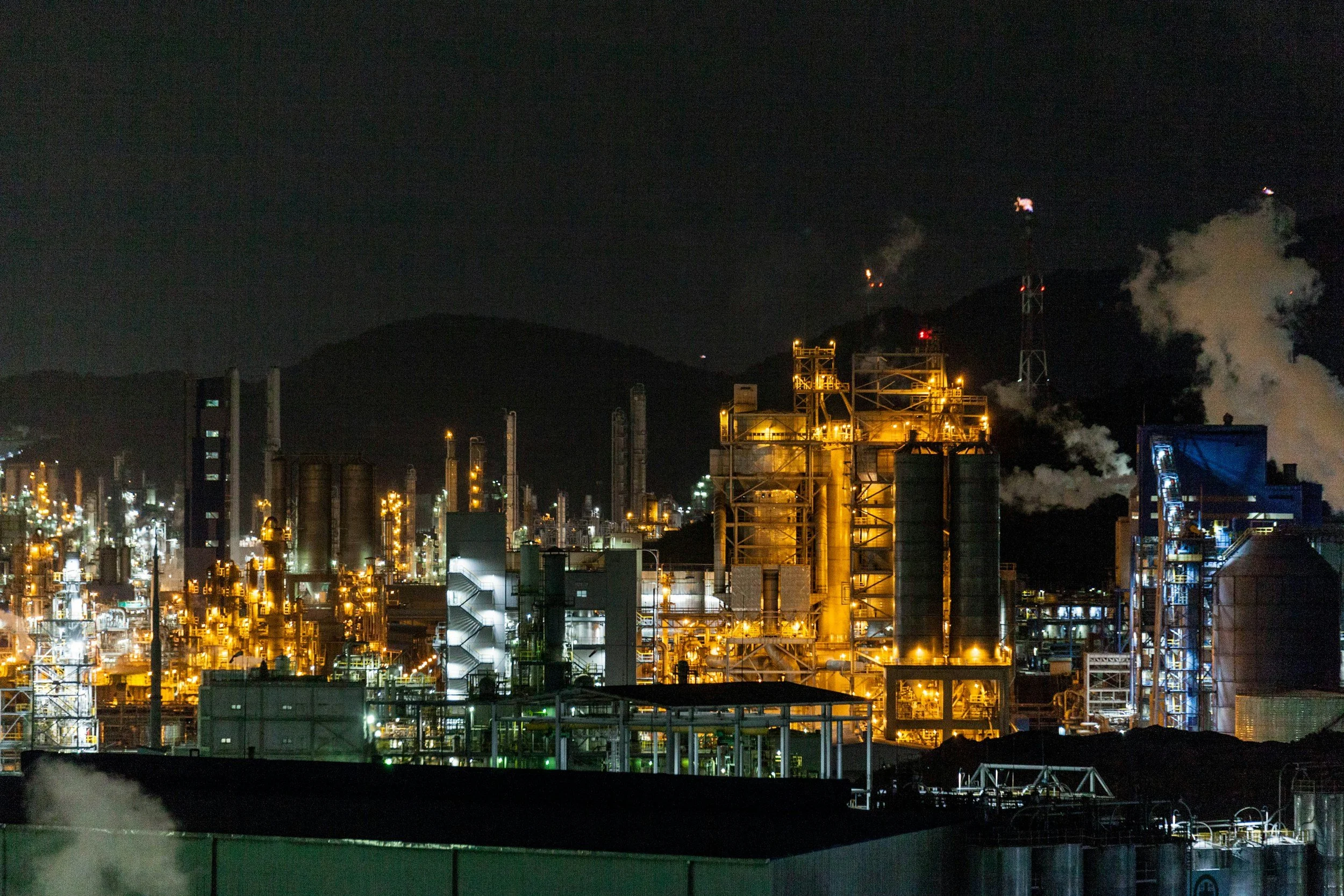 Nighttime view of an industrial factory or refinery with illuminated structures and smoke or steam emissions, set against dark mountains.