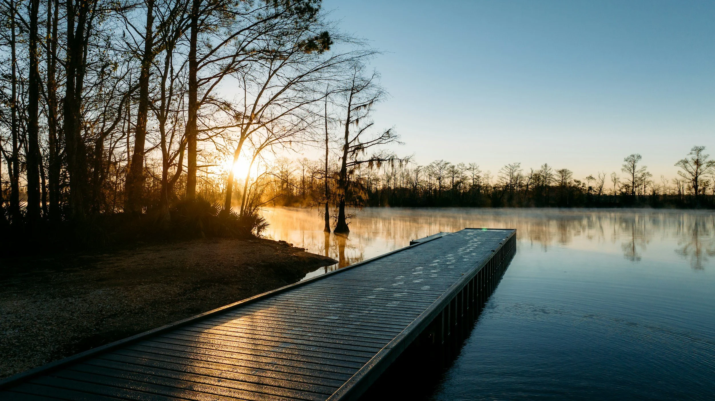 A wooden dock extends over calm water near a forested shoreline during sunrise, with mist rising from the water and trees silhouetted against a clear sky.