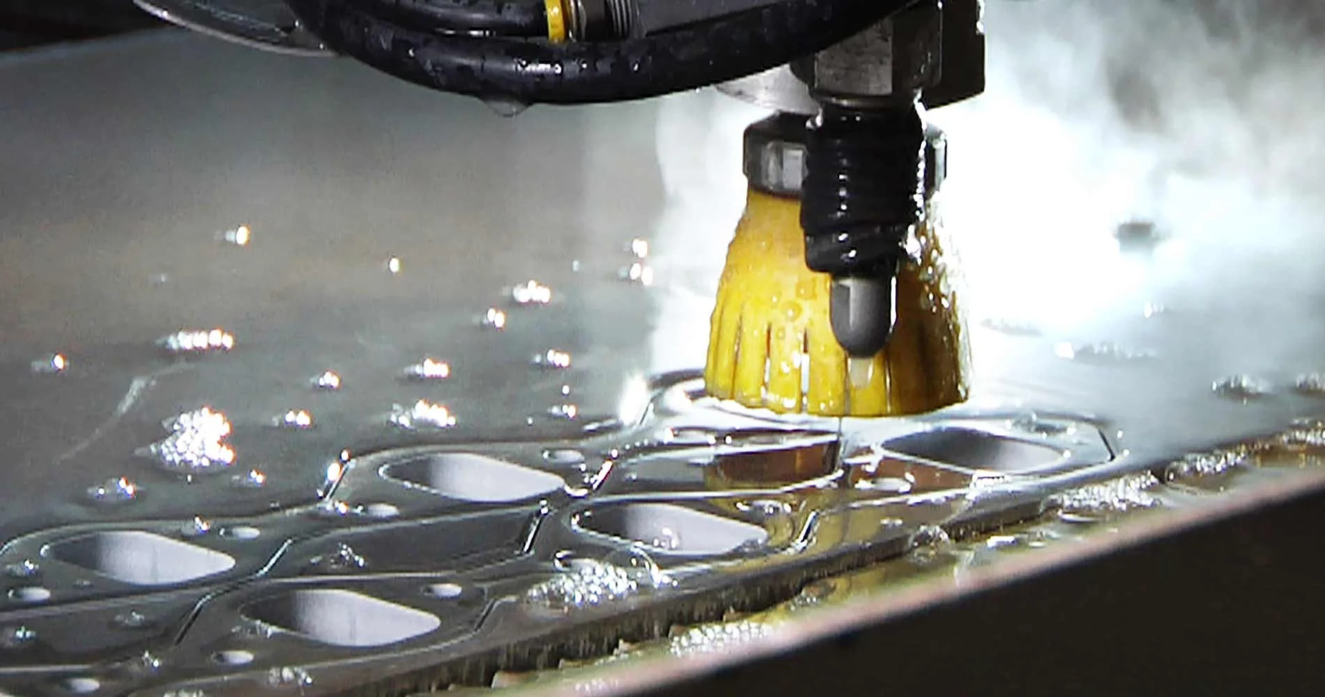 Close-up of a robotic cleaning tool with a yellow scrubber, cleaning a metallic surface with multiple holes and bubbles of cleaning liquid.