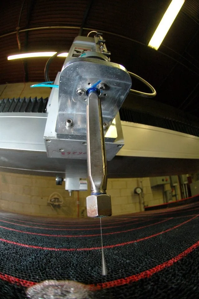 Close-up of a precision manufacturing machine with a metal arm and a thin string or filament hanging down, with a black textured surface below it.