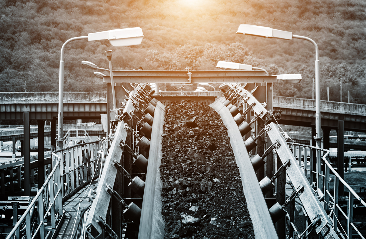 Industrial conveyor system transporting raw coal, with hillside and bridge in background.