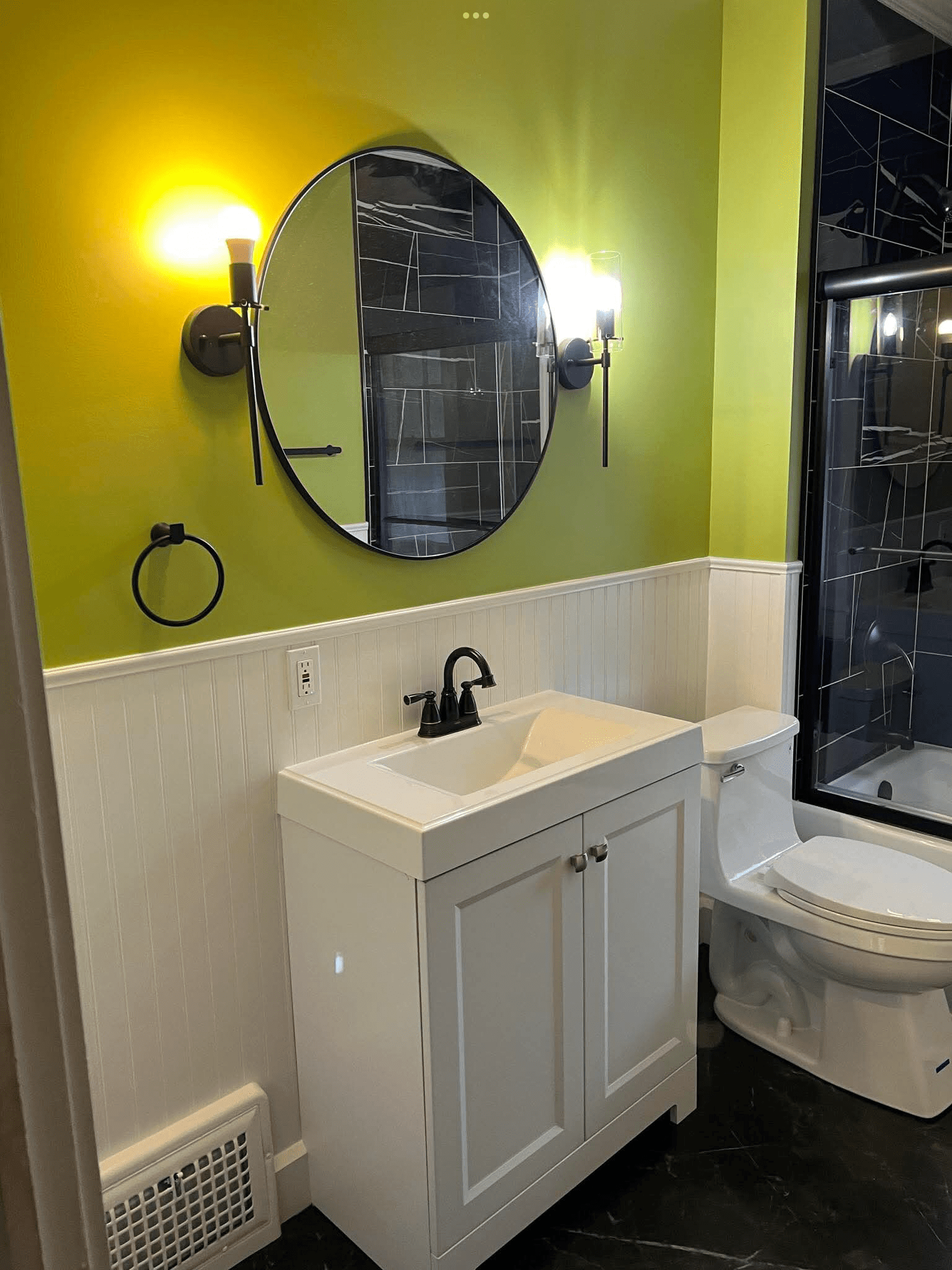 White bathroom vanity with black faucet, round mirror, wall sconces, and custom beadboard walls in a remodeled Northeast Ohio bathroom.